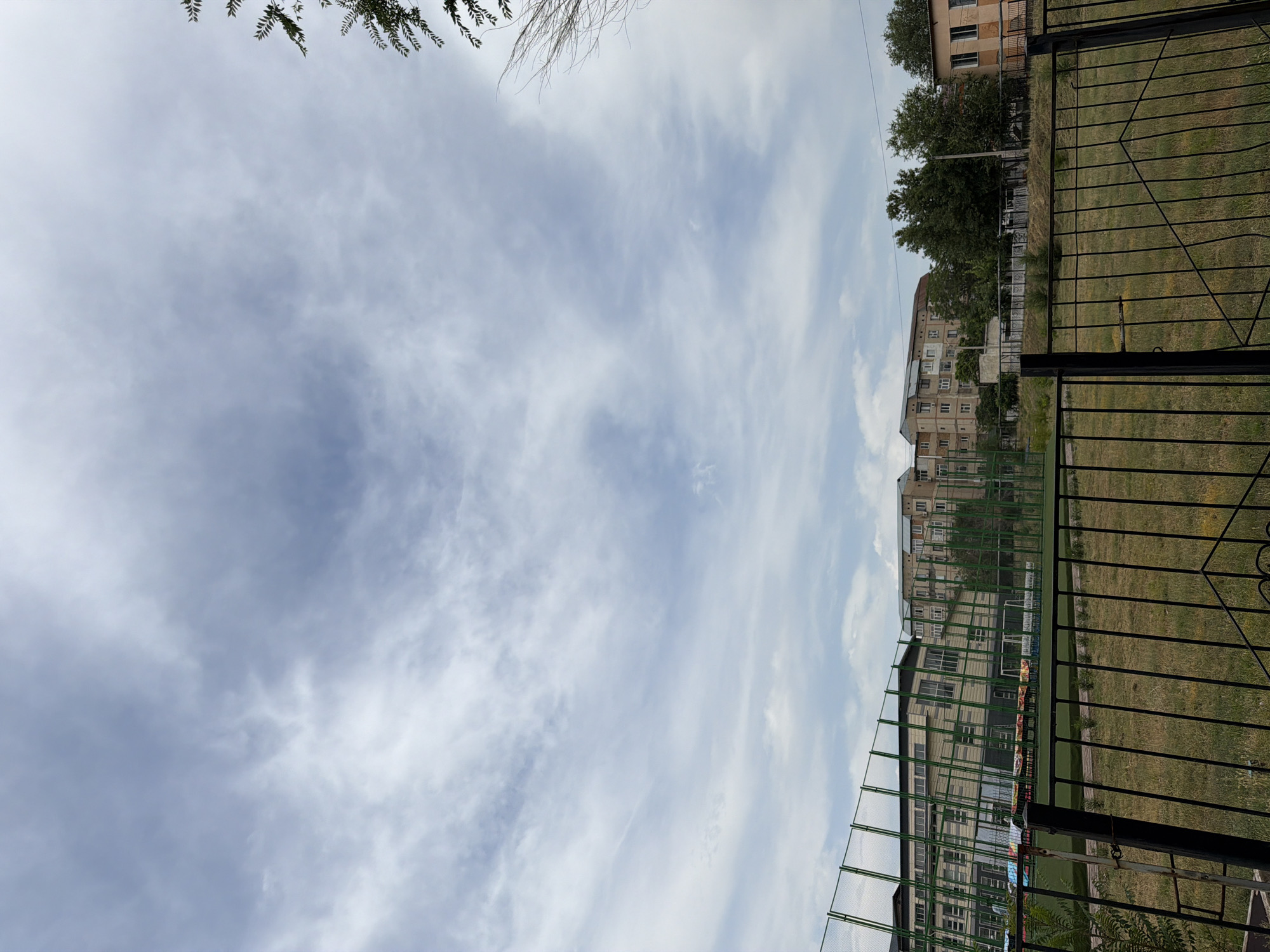 The image depicts a view of a schoolyard or sports field in Shymkent, Kazakhstan. In the foreground, a black metal fence runs horizontally across the frame, with a patch of dry grass and weeds behind it. Beyond the fence, a green sports field with what appears to be artificial turf is visible. Along the far edge of the field, a high green mesh fence encloses the area. Behind this fence, a multi-story building with a light-colored facade and many windows is visible. To the right of this building, there are several trees, and further to the right, another building with a light orange facade and windows is present. In the upper portion of the image, a vast sky filled with scattered clouds is depicted. The overall scene suggests an outdoor area of an educational institution or a sports facility.