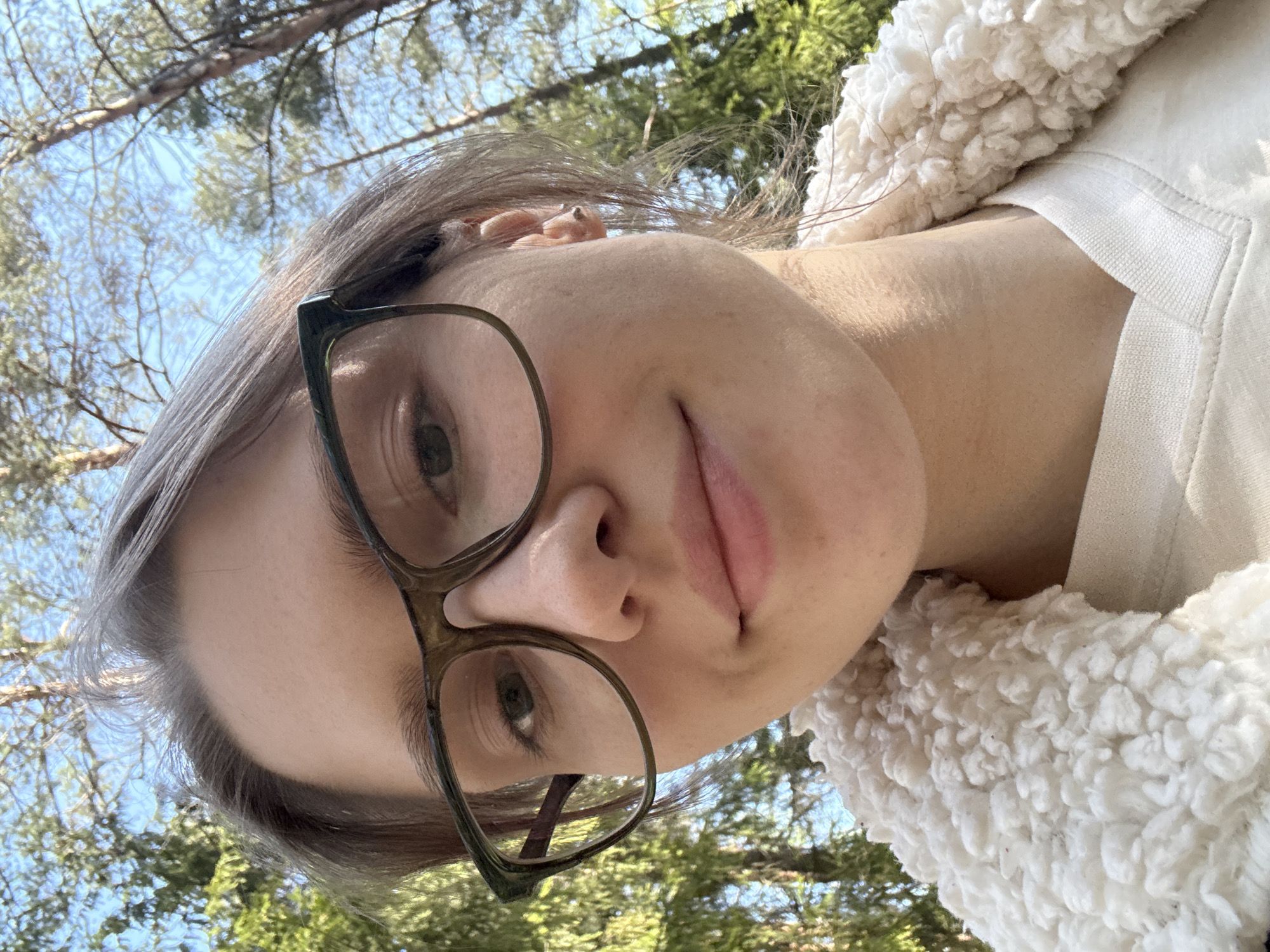 A person with light skin, short gray hair, and wearing glasses is the primary subject of the image. The person is looking slightly upwards towards the camera. The person is wearing a light-colored fluffy jacket or sweater. The background shows green trees and a blue sky, suggesting an outdoor setting. The location is Solnechnoye, Russia.
