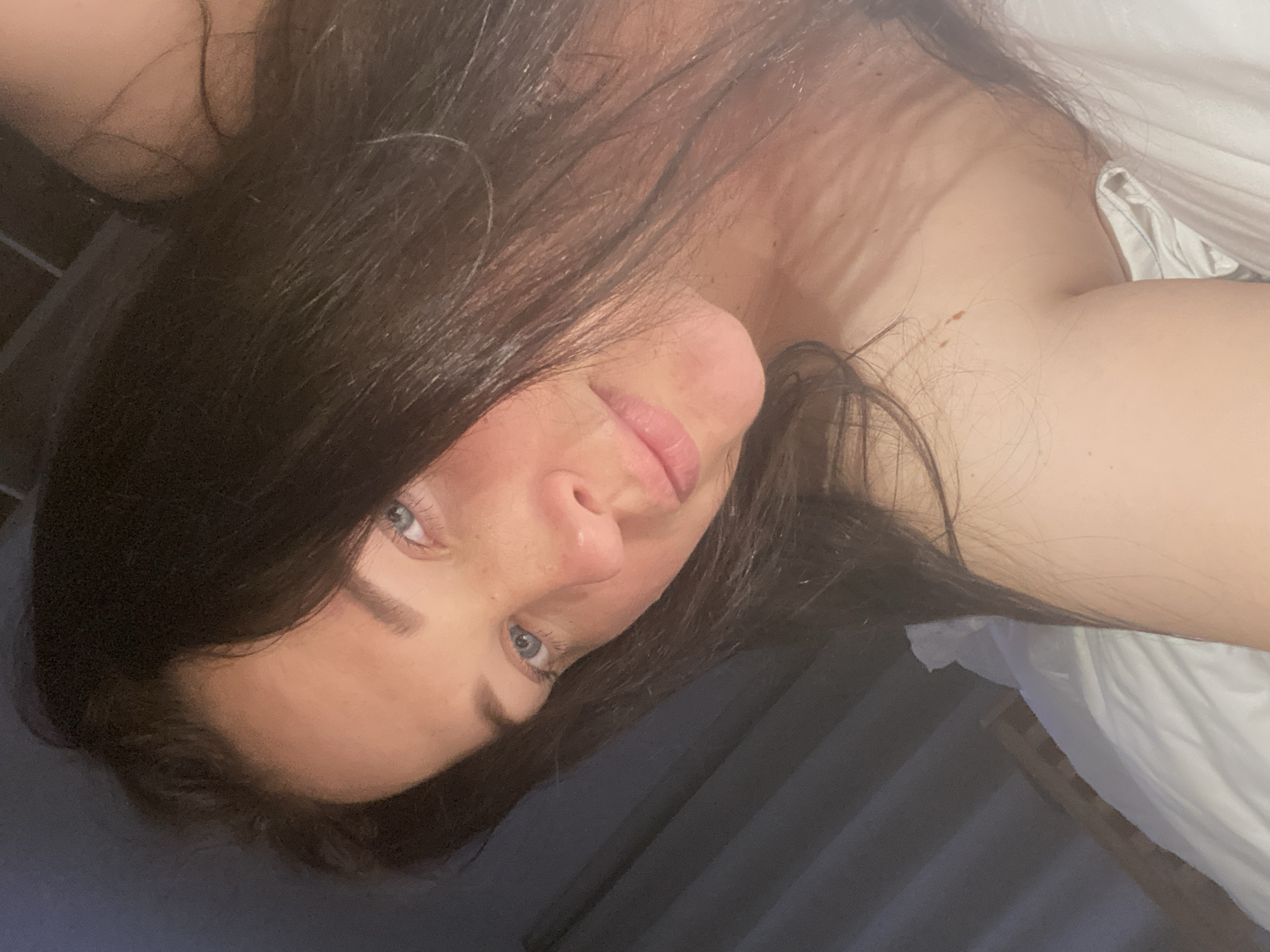 A woman with long dark hair and light eyes is lying down. She is wearing a white top or is lying on white bedding. The background appears to be a combination of dark blue fabric or bedding and what may be a headboard or wall. It is not possible to determine if the location is Sochi, Russia, or any other city based on the provided image.
