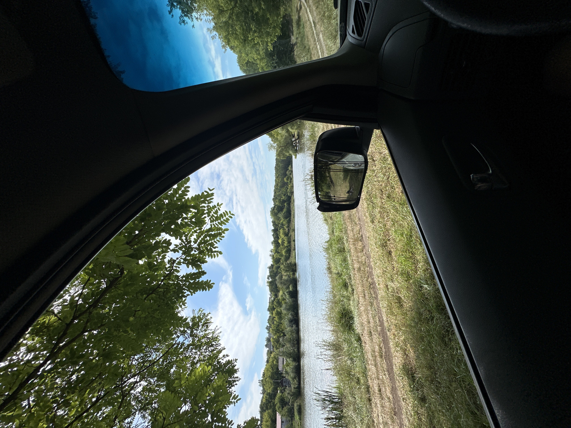 The image is taken from inside a vehicle, looking out the side window. The view is of a serene natural landscape featuring a body of water, possibly a river or lake, with lush green trees and vegetation lining its banks. The sky is a vibrant blue with scattered white clouds, suggesting a clear day. In the foreground, the interior of the car is visible, including the door panel, a side mirror reflecting the scenery, and a portion of the steering wheel. The overall impression is one of a peaceful rural setting, likely outside of a major urban area. Based on the provided information, this scene is located in Makiv, Ukraine, a village situated in the Cherkasy Oblast.