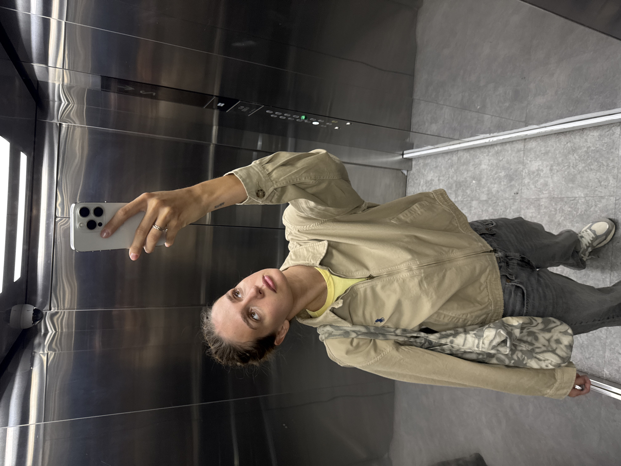 A person is inside an elevator, taking a selfie with a smartphone. The elevator has stainless steel walls and a control panel with illuminated buttons. The floor is gray. The person is wearing a beige jacket over a yellow shirt, gray jeans, and sneakers. They also have a patterned shoulder bag.