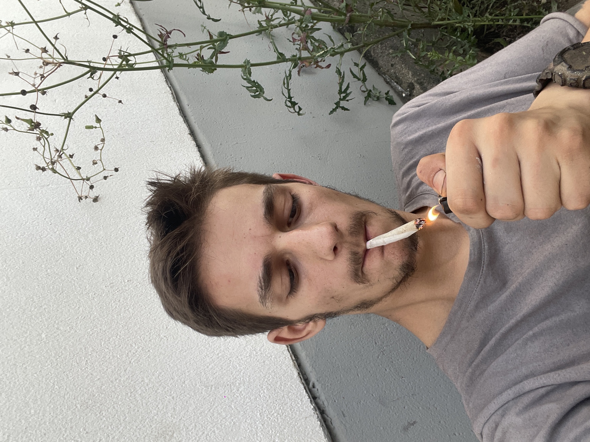 A close-up, waist-up shot shows a young person with short, brown hair and a light mustache and beard, lighting a cigarette with a black lighter. The person is wearing a grey t-shirt and has a dark-colored watch on their left wrist. They are looking down at the cigarette, which is held between their lips. The background consists of a textured white wall, a grey concrete surface, and some greenery. The lighting is natural, and the image is taken from a low angle.