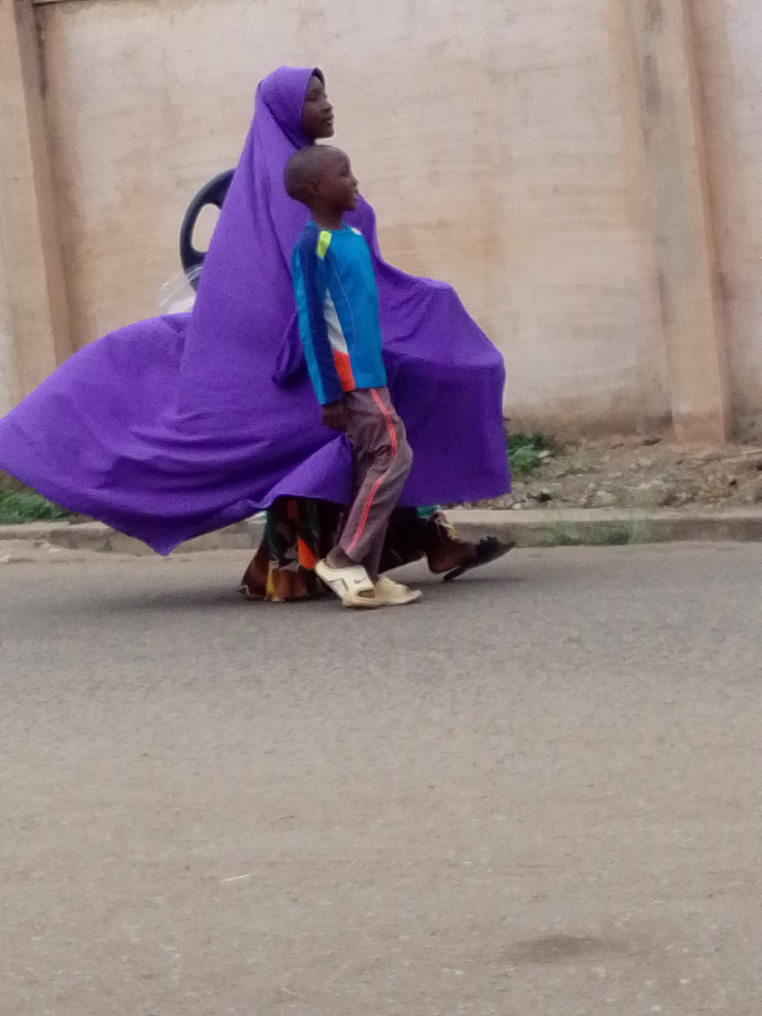 An outdoor, eye-level shot captures two individuals, an adult and a child, walking alongside a light-colored wall. The adult, positioned behind the child, wears a striking purple head covering and a voluminous, flowing purple garment that drapes around her. A dark, possibly curved object is visible behind her, partially covered by the fabric. Her lower legs and feet, clad in dark sandals, are visible beneath the flowing attire. The child, a young boy, walks slightly ahead and to the left of the adult. He is dressed in a blue long-sleeved shirt with accents of orange and yellow, paired with brown pants featuring red stripes down the sides. He wears light-colored sandals.
Both individuals are in profile, looking towards the left side of the frame. They appear to be walking on a paved surface, possibly a road or sidewalk, with a narrow strip of dirt and sparse vegetation visible between the pavement and the background wall. The wall itself is a plain, light beige or tan color. The lighting suggests daytime, possibly overcast or during the softer hours of morning or late afternoon, as there are no harsh shadows. No text is visible in the image. The scene conveys a sense of everyday movement in an unidentified outdoor setting.