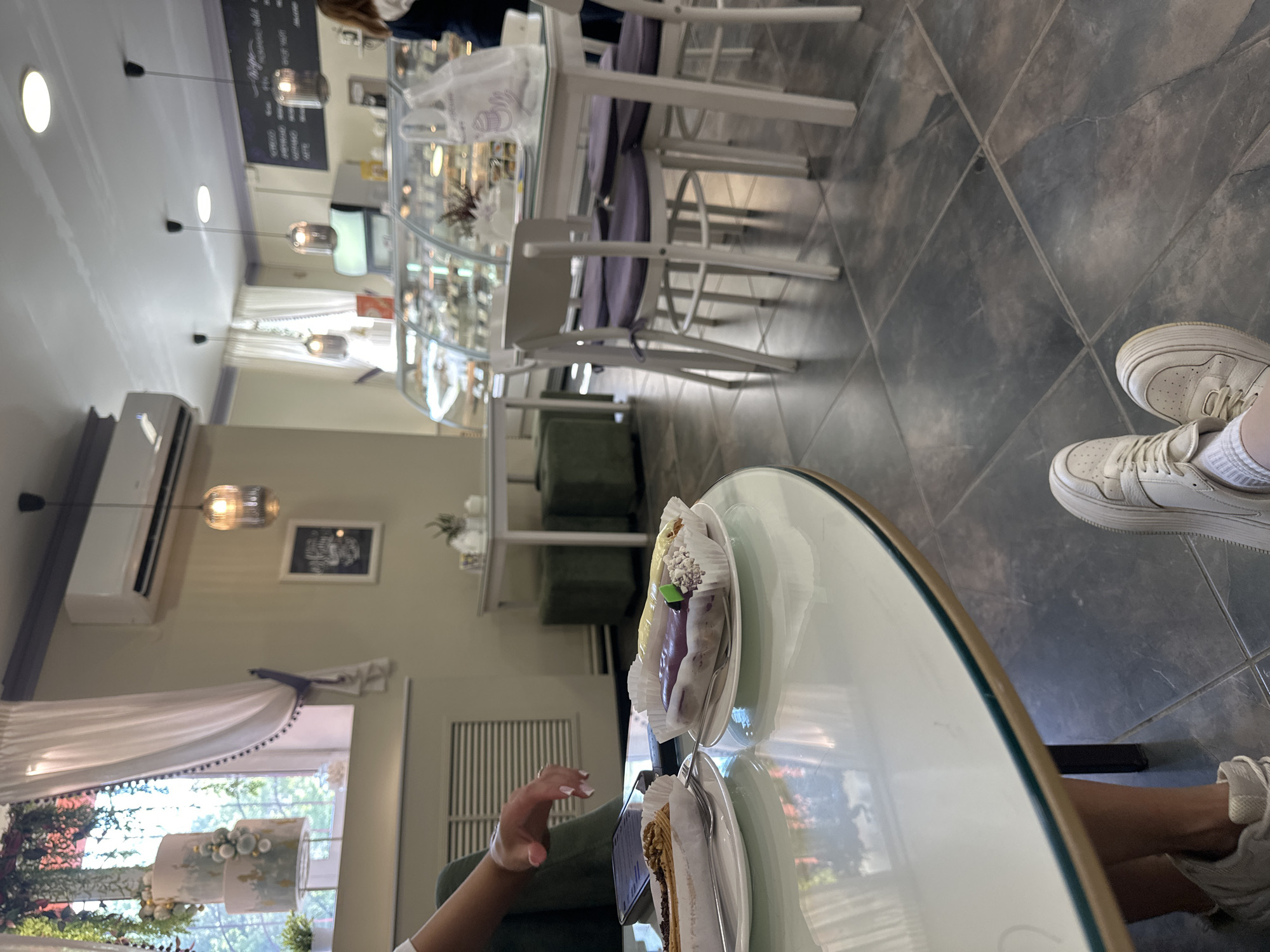 The image depicts the interior of a cafe. In the foreground, a person's legs and feet, clad in white sneakers and socks, are visible. They are seated at a round, glossy table where two eclairs on white paper wrappers rest on white plates with forks. To the left, a person's arm and hand are seen reaching towards a smartphone on the table. In the background, several tables and chairs are arranged, with one white table featuring purple cushions on its chairs. A display case filled with pastries is visible behind the tables. A three-tiered cake is displayed on a stand near a window with white curtains. The cafe appears to have a modern, clean aesthetic with tiled flooring. There is no explicit indication of the city of Perm, Russia, in the image.