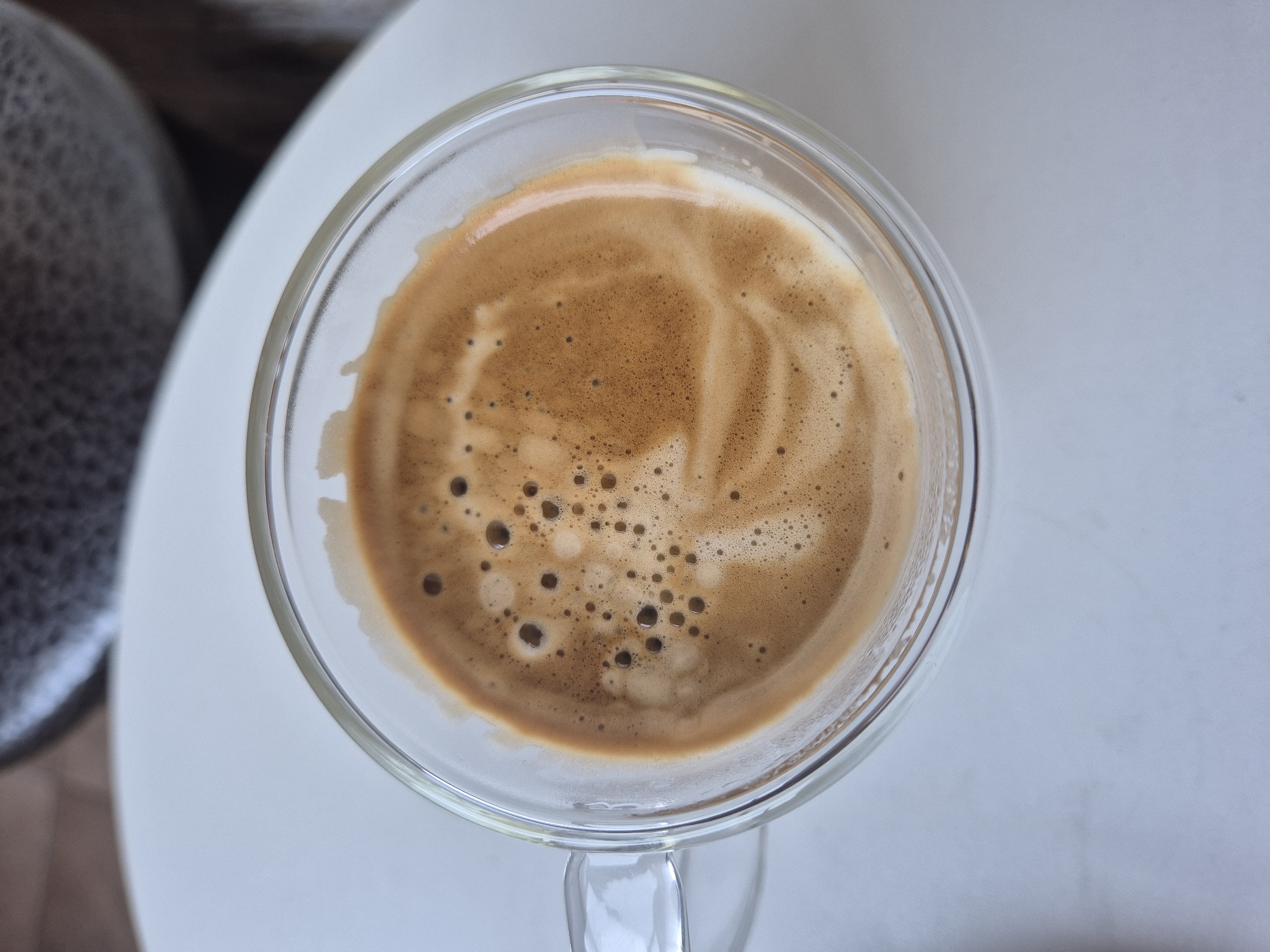 A close-up, overhead shot shows a clear glass mug filled with espresso. The coffee has a rich, foamy crema with a swirl pattern and scattered bubbles. The mug is on a white surface, with a blurred background that appears to be a table or countertop, and possibly a textured object in the upper background. The lighting suggests an indoor setting.