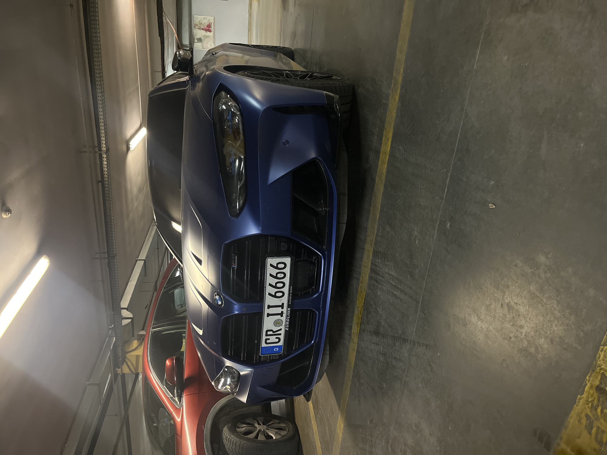 The image depicts the front of a dark blue BMW car parked in an indoor parking garage. Adjacent to the BMW is a red vehicle, partially visible. The BMW has a European license plate that reads "CR II 6666" with a German “D” country code, indicating the car is registered in Germany. The surroundings suggest a typical indoor parking environment, with concrete floors marked by yellow parking lines. The lighting is provided by fluorescent overhead lights, casting shadows within the space. There is no indication of the image's location within Varna, Bulgaria.