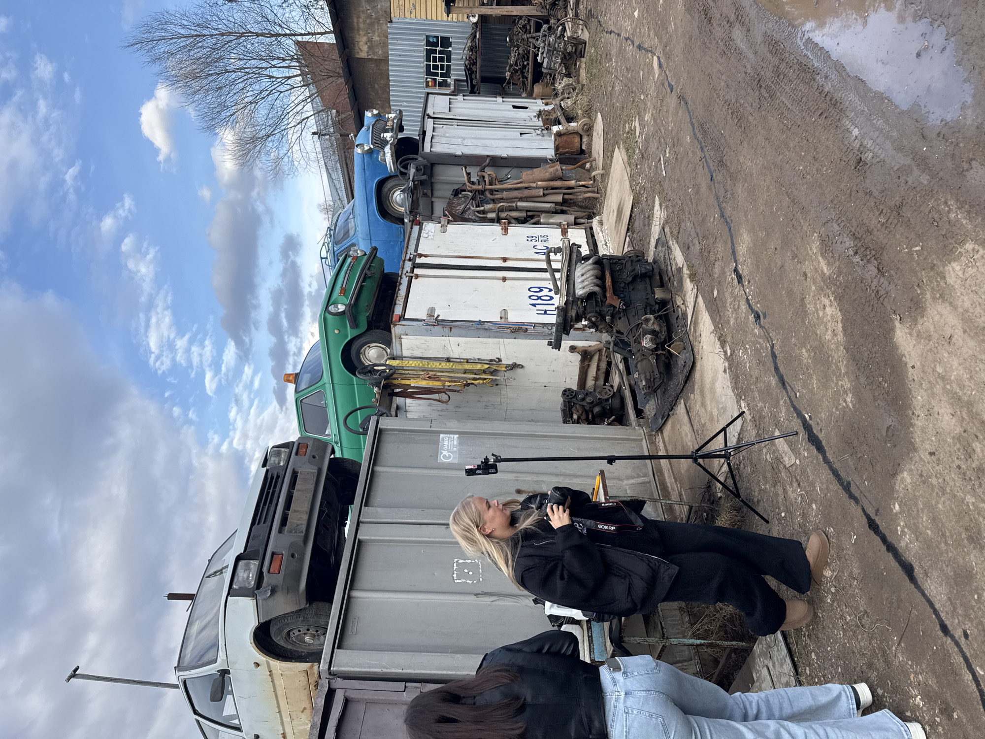 The image depicts an outdoor scene at what appears to be a car junkyard or auto dismantling facility in Chaikovsky, Russia. The sky is overcast with clouds, suggesting daytime, and the ground is muddy with tire tracks and puddles, indicating recent rain.
Two women are present. In the foreground, a blonde-haired woman, dressed in a black jacket and black pants, is actively holding a Canon EOS RP camera to her eye, engaged in photography or videography. A black tripod with a phone mount stands nearby. Behind her, another woman is partially visible, wearing a black jacket and light blue jeans.
The site is filled with numerous old vehicles and discarded car parts. Several cars, including a white van (resembling a UAZ-452), a green car (similar to a Lada/Zhiguli), and a blue sedan (like a GAZ-21 Volga), are stacked on top of large corrugated metal containers, which also serve as storage units. Piles of car engines, exhaust pipes, and various metal components are scattered across the ground and on containers. In the background, there are buildings with corrugated metal siding and roofs, along with a bare-branched tree.
Visible text includes "CANON EOS RP" on the camera. On a white container, partially visible markings include "H189", "59 RUS", and "4C". The word "DIABLO" can be faintly seen on a grey container. The overall scene suggests a creative project or media shoot taking place within this industrial and cluttered environment.