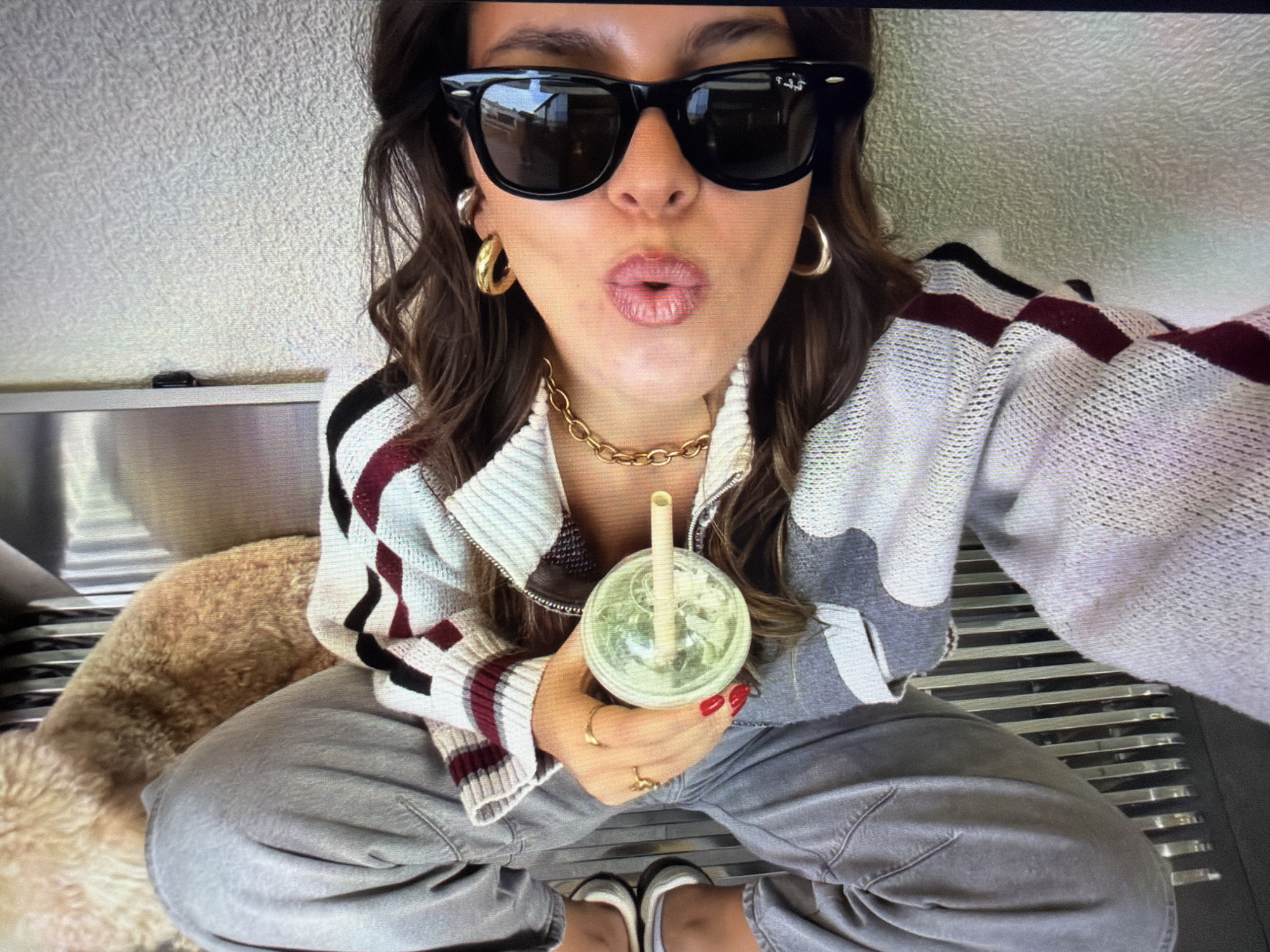 This high-angle selfie captures a person, likely female, sitting indoors on a metal slatted bench in what appears to be Ros Na Rí, Ireland. The subject is seen from the chest up, with her legs and feet also visible. She has dark, wavy hair and is wearing black sunglasses, a thick gold chain necklace, and prominent gold hoop earrings. Her lips are puckered in a playful or kissing expression, enhanced by a light-colored lip gloss.
She is dressed in a zip-up knit sweater or jacket featuring a distinctive pattern of light grey, white, black, and maroon stripes and abstract shapes. Her fingernails are painted a vibrant red, and she wears multiple gold rings on her fingers. In her right hand, she holds a transparent plastic cup containing a green beverage, possibly a smoothie or matcha, with a light-colored straw.
To her left, partially visible, is a fluffy, light brown animal, likely a dog, resting beside her on the bench. She is wearing loose-fitting grey pants and two-toned flat shoes, possibly white and black. The background includes a textured, light-colored wall. The brand name "Ray-Ban" is visible on the temple of her sunglasses. The image conveys a casual and relaxed atmosphere.