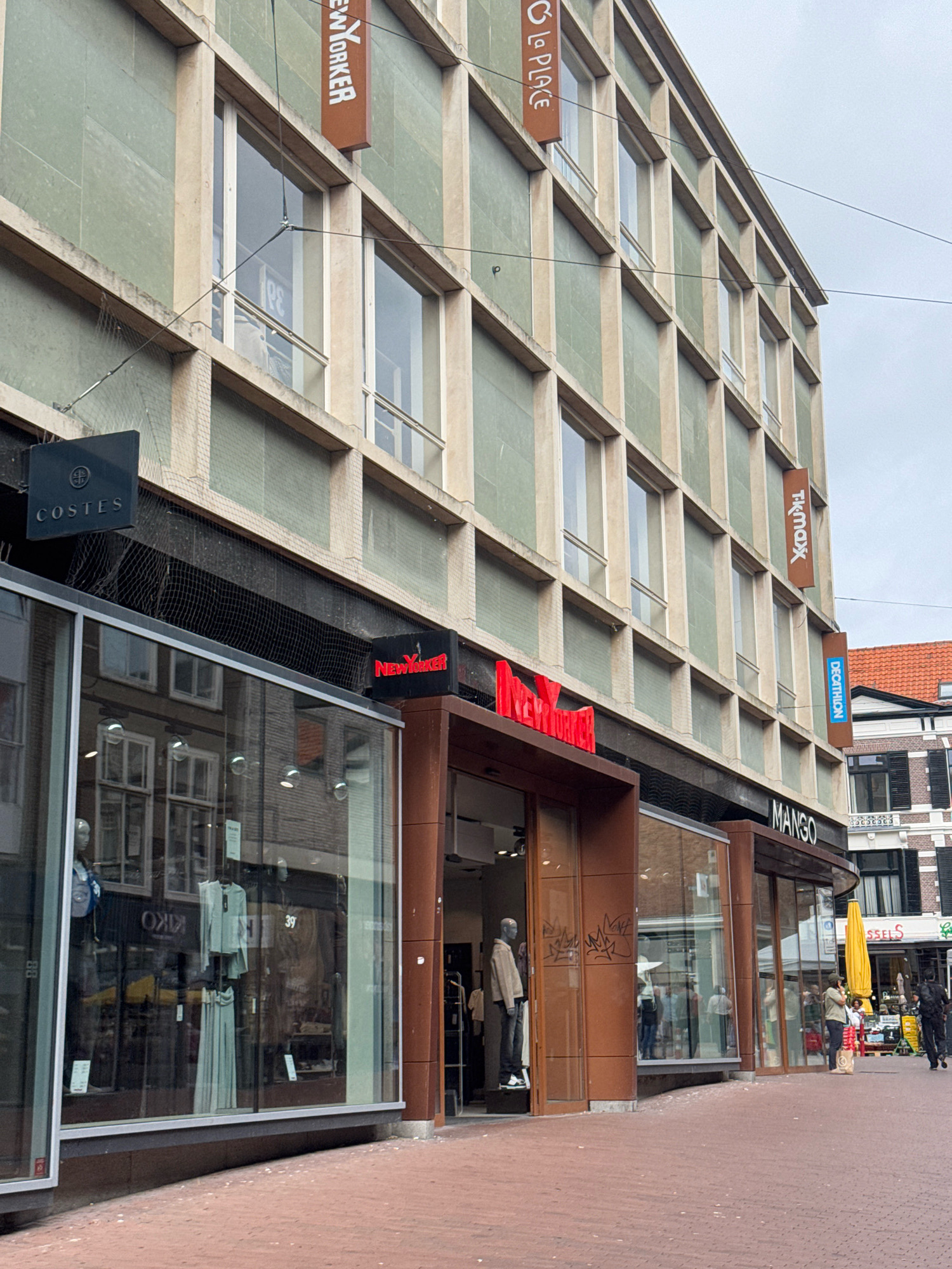 This is a medium shot of a street scene in Nijmegen, Netherlands, focusing on a commercial building with several storefronts. The building has a facade of light-colored concrete pillars and pale green panels, with multiple floors of windows. Several retail stores are visible at ground level.
Prominently displayed signage includes "NEW YORKER" in red neon letters above one entrance, and "MANGO" in white lettering on a black background above another. Above the "NEW YORKER" store, there is a dark blue sign with white text reading "COSTES." Further up the building facade, vertical signs indicate "NEW YORKER," "LA PLACE," "TK MAXX," and "DECATHLON."
The storefronts feature large glass windows, showcasing merchandise inside. One window displays mannequins dressed in clothing and a selection of apparel. Another window shows reflections of buildings and people across the street. The street itself is paved with reddish-brown brickwork.
In the foreground, on the right side of the image, several people are walking along the sidewalk. One person is holding a yellow umbrella. The sky is overcast. The overall impression is of a typical urban shopping street.