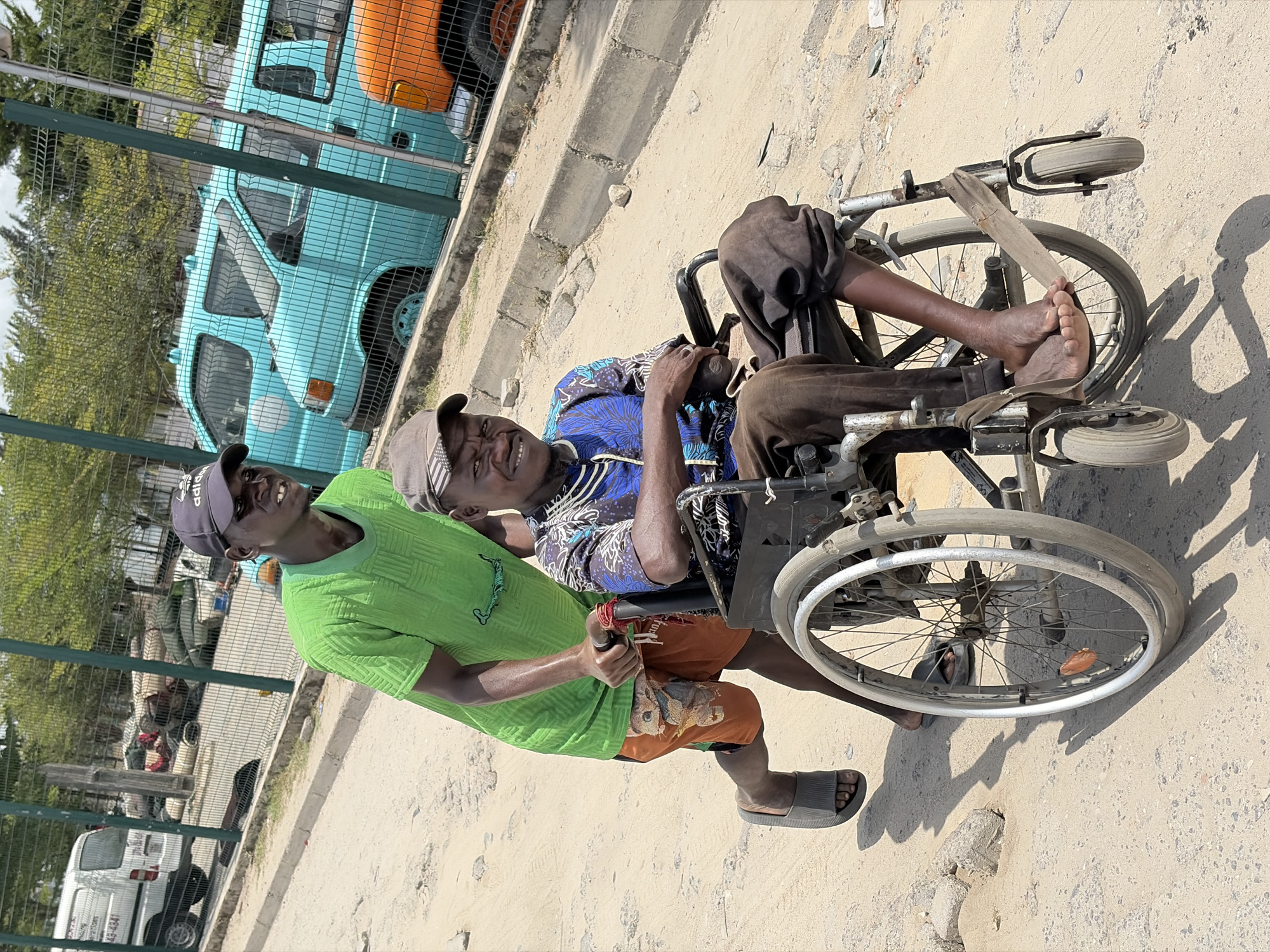 The image depicts two Black men in Igboefon, Nigeria. One man, wearing a green Adidas t-shirt, a gray cap, and orange shorts, is pushing the other in a wheelchair. The man in the wheelchair wears a blue patterned shirt and brown pants. He is barefoot, and a piece of wood is positioned above his feet within the chair. They are on a dirt and concrete road next to a fence. Behind the fence, there are several cars and vans visible, including a light blue van.