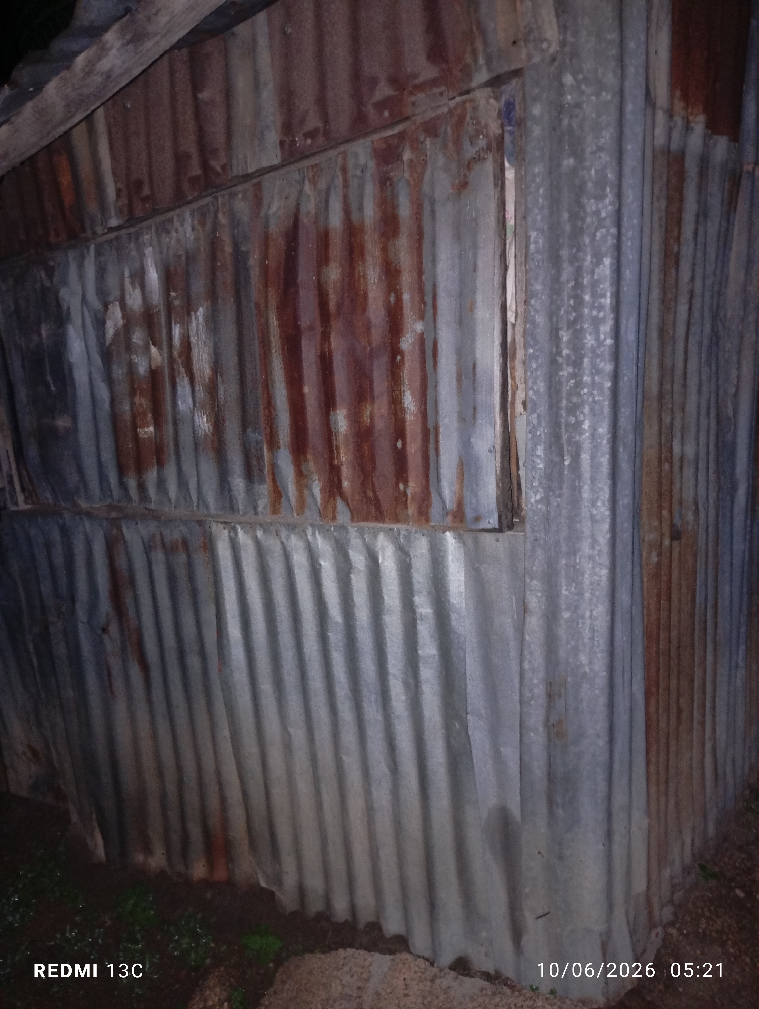 A weathered, corrugated metal structure, possibly a makeshift shelter or shed, stands in an outdoor environment. The metal sheeting shows signs of rust and age, with some sections appearing more oxidized and reddish-brown than others. The structure has a peaked roofline partially visible at the top left, supported by wooden beams. There appears to be a narrow opening or window in the upper section of the wall. The ground surrounding the structure is covered in a mixture of soil, gravel, and some sparse vegetation. A timestamp in the bottom right corner indicates the image was taken on "10/06/2026 05:21". The bottom left corner displays "REDMI 13C". The scene is dimly lit, suggesting it might be early morning or late evening. The specific location in Jos, Nigeria, cannot be determined from the visual information provided.