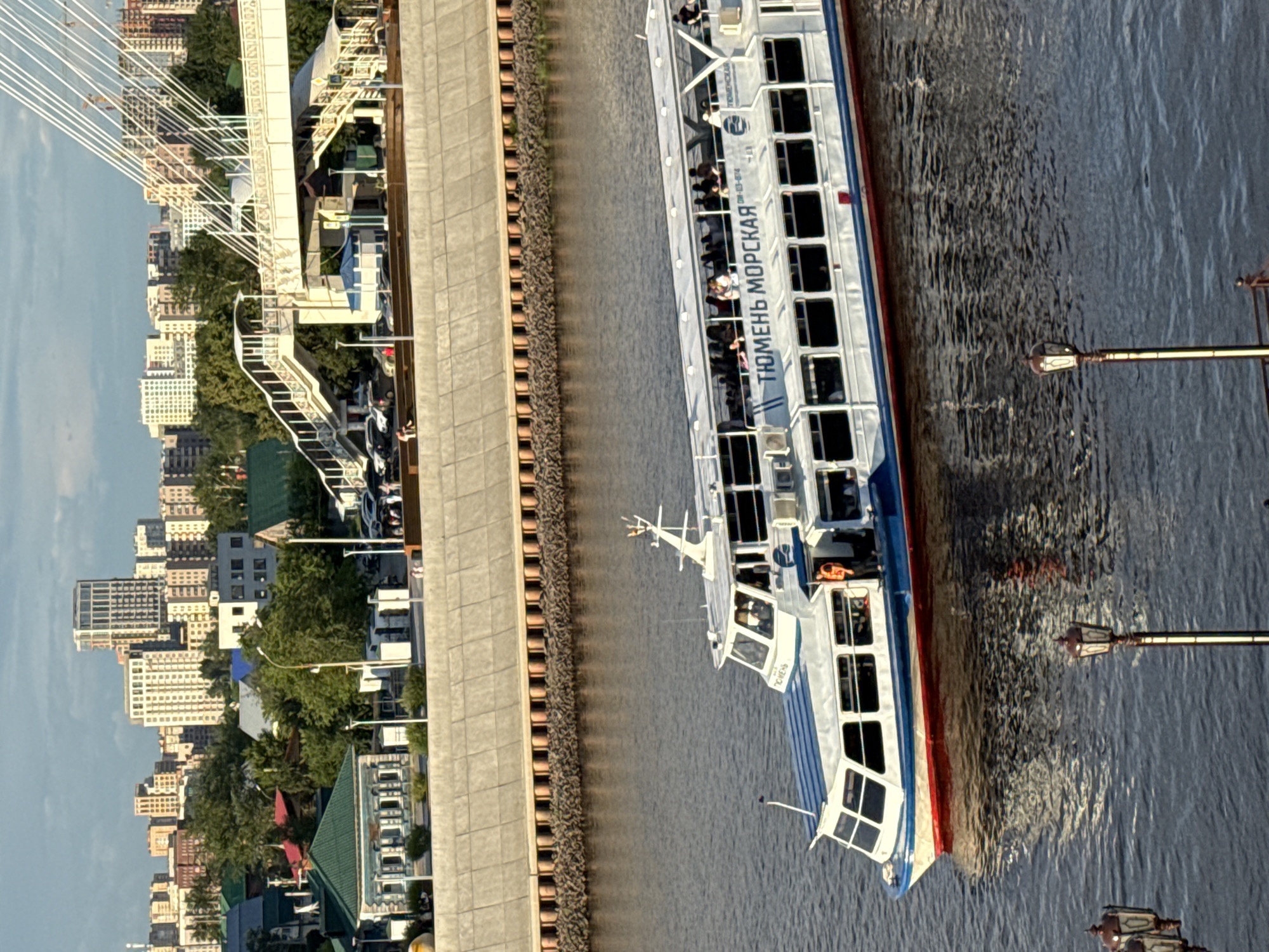 The image shows a boat on the Tura River in Tyumen, Russia. The boat is white with blue and red accents and has "ТЮМЕНЬ МОРСКАЯ" written on its side. There are many passengers on the boat, visible through the windows and on the upper deck. In the background, the cityscape of Tyumen is visible, featuring modern buildings, older structures, and lush green trees. A pedestrian bridge with cables is also prominent in the background. The river is calm, reflecting the sky and the surrounding structures. The lighting suggests it is late afternoon or early evening. Several lampposts are visible in the foreground, indicating a public walkway or promenade.