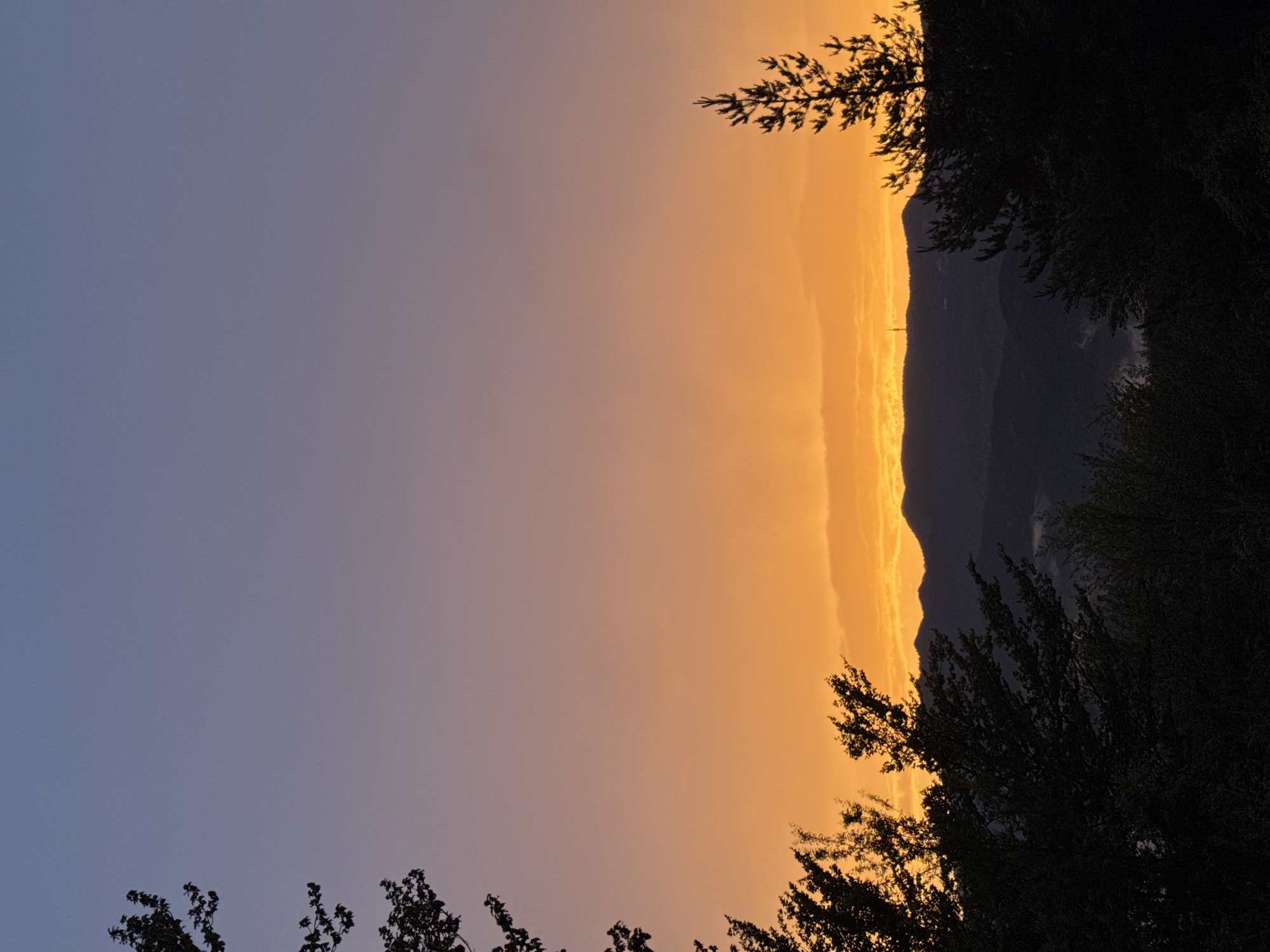 This image captures a sunset over the city of Strazha, Bulgaria. The sky transitions from a deep blue at the top to a vibrant orange and yellow near the horizon. Silhouetted trees frame the scene on the left and right sides. In the background, a range of dark, rolling hills and mountains is visible, with a prominent communication tower standing atop one of the peaks. Wisps of mist or low clouds can be seen in the valleys between the mountains. The overall impression is one of natural beauty and tranquility as day turns to night.