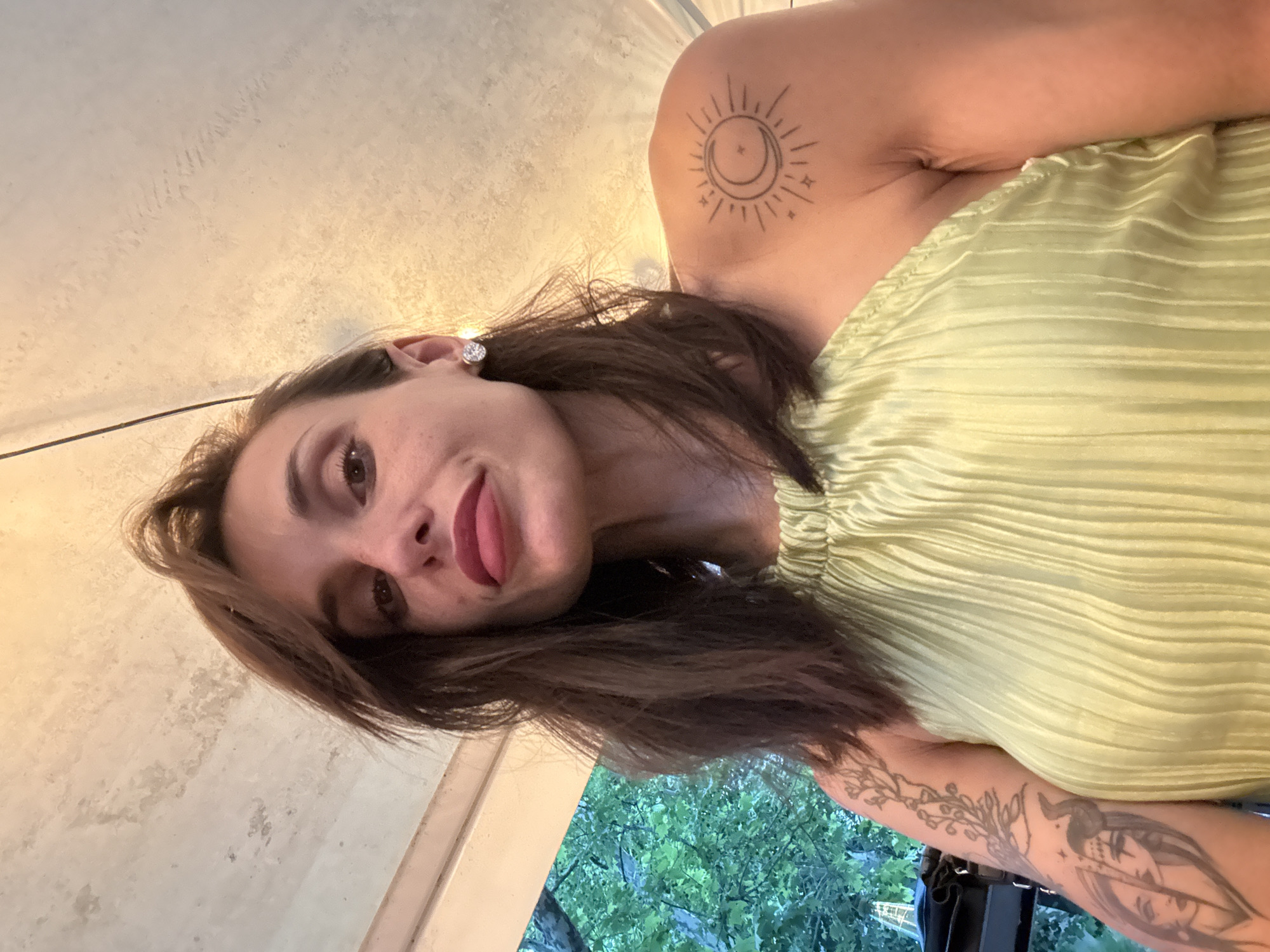 A woman with brown hair and dark eyes is pictured from a low angle, looking up at the camera. She is wearing a light green halter top and large, sparkling earrings. She has tattoos on her left arm and right shoulder. The background shows trees and a ceiling. The image was taken in Pleven, Bulgaria.