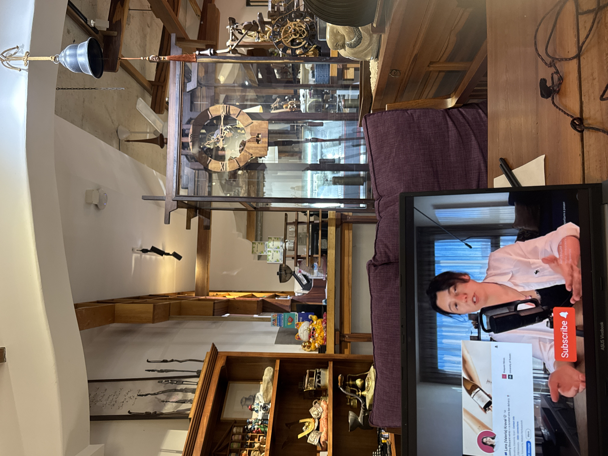 The image depicts an indoor scene, likely a shop or a cafe, in Nha Trang Ward, Vietnam. In the foreground, a laptop displays a video call or recording. A woman with dark hair is speaking to the camera, gesturing with her hands. A "Subscribe" button is visible on the laptop screen.
The background reveals a richly decorated interior with wooden shelves filled with various items, including figurines, bottles, and what appear to be antique artifacts like a large clock with exposed gears. A plush purple sofa is positioned behind the laptop. The ceiling has exposed wooden beams and architectural details. A bell hangs from the ceiling, adding to the eclectic decor. The overall impression is a well-curated space with a mix of traditional and possibly vintage items.
