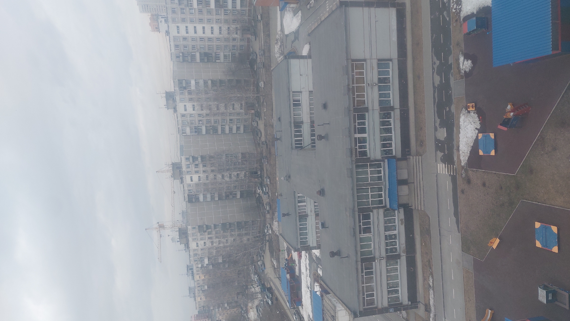 The image depicts an aerial view of a residential area in Novosibirsk, Russia, characterized by a mix of high-rise apartment buildings and lower-rise industrial or commercial structures. The apartment buildings are densely packed, showcasing a uniform gray facade with rows of windows. A construction crane is visible among the buildings. Adjacent to the residential area is a long, gray building with multiple windows, likely a school or factory, running parallel to a road with some snow accumulation. A playground with colorful play structures sits next to this building. A few scattered cars are parked along the streets. The overall atmosphere suggests a cold, possibly overcast day.