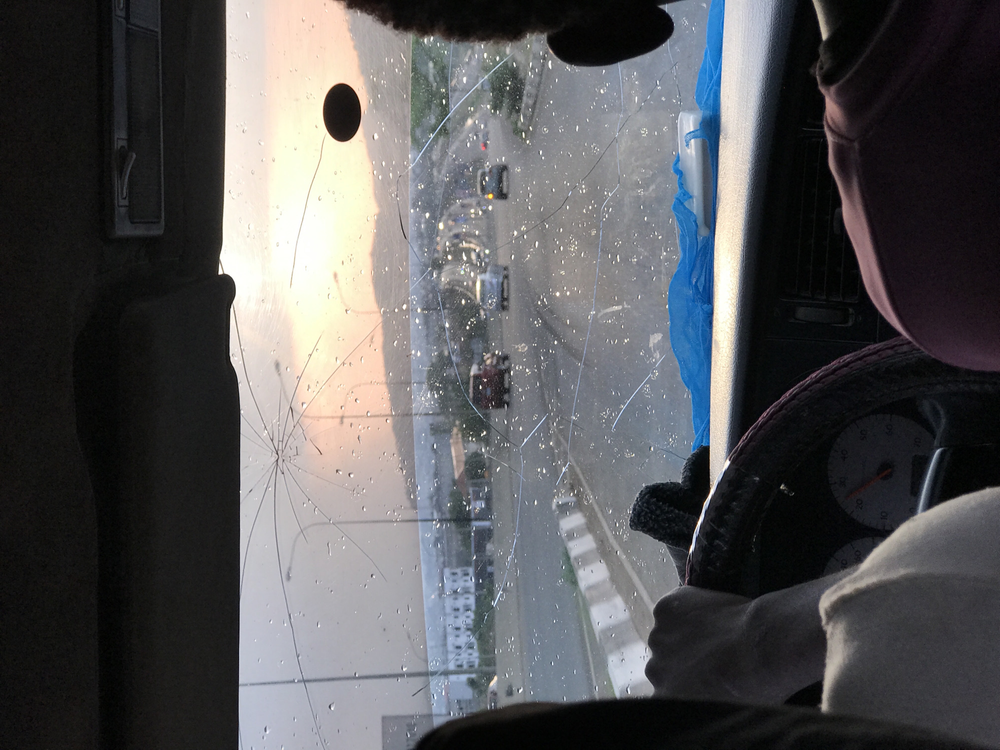 The image is taken from inside a vehicle with a severely cracked windshield. The vehicle appears to be stationary, facing an urban street. Raindrops are visible on the glass. Through the damaged windshield, one can see cars on the road, buildings, streetlights, and a sky with an orange hue, suggesting either sunrise or sunset. A steering wheel is in the foreground, with a person's hands gripping it. A speedometer is partially visible. To the left of the windshield is a dark-colored interior panel of the vehicle with a metallic fixture. To the right is another section of the dashboard, accompanied by a piece of blue fabric. The location is indicated as Jiru, Nigeria.
