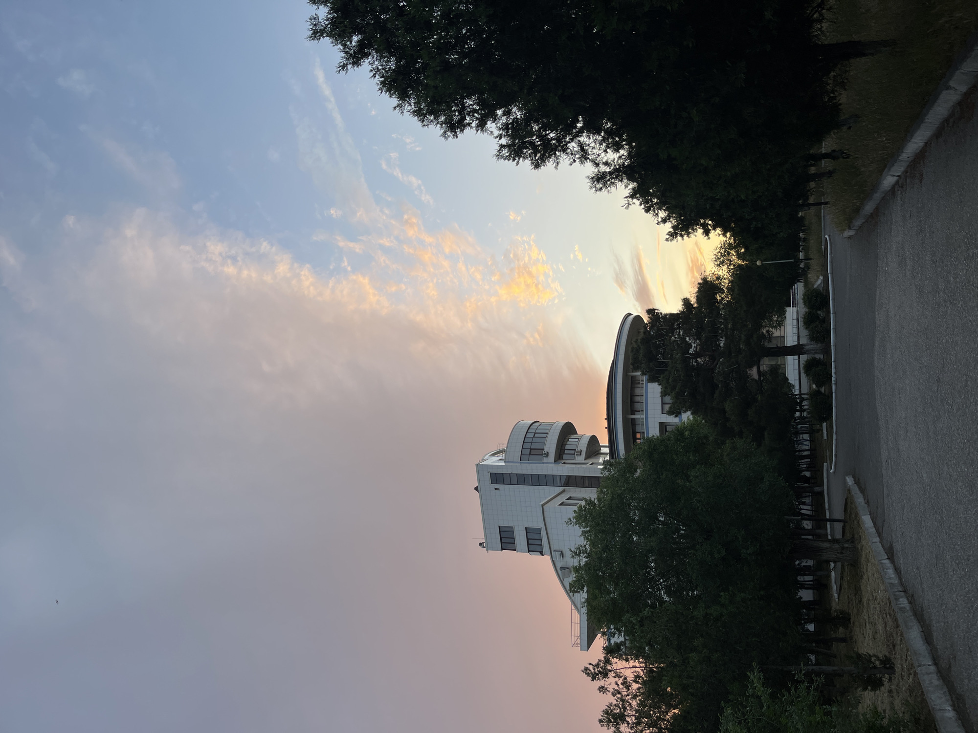 The image shows a white building with a curved roofline in Kaspiysk, Russia, likely photographed in the late afternoon or early evening. The sky is multi-toned, ranging from pinkish-purple to blue with hints of yellow where the sun might be setting. Trees frame the top of the building and line the side of the road to the right of the building. The road is grey and has a concrete curb separating it from the dry grassy verge. A streetlight is visible. The architectural style of the building appears to be modern.