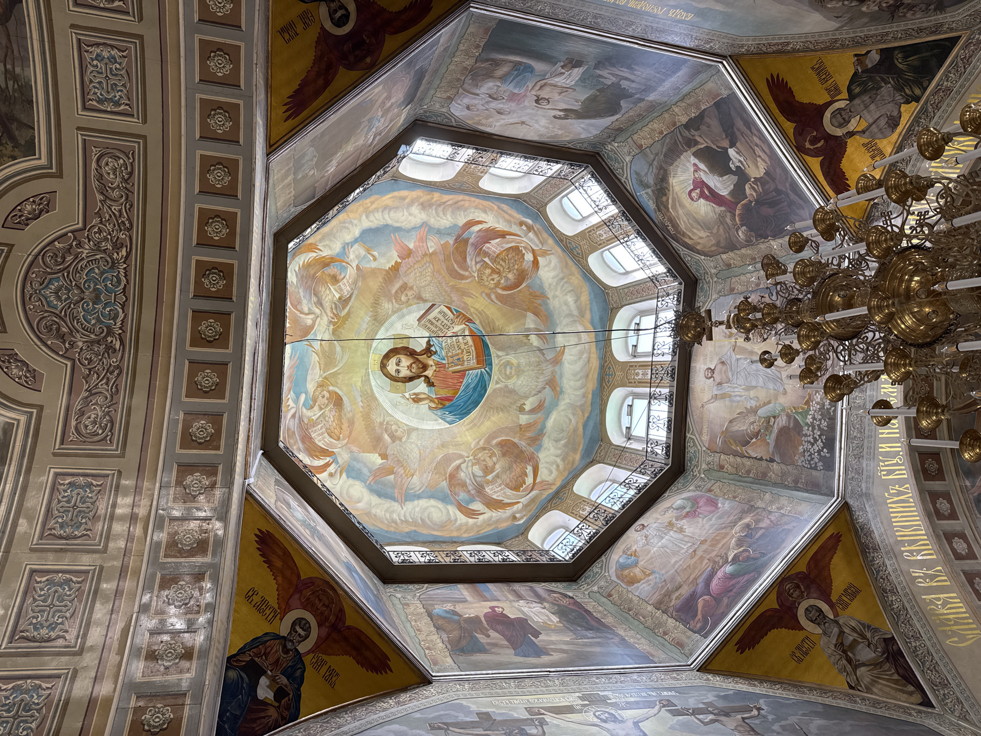 The interior of a grand Orthodox church or cathedral is captured from a low, upward-looking perspective, showcasing its ornate domed ceiling and walls adorned with numerous religious murals and decorative elements.
The central feature is a large, octagonal dome, at the apex of which is a vibrant painting of Jesus Christ holding an open book, surrounded by angels floating on clouds against a blue and yellow sky. The base of the dome features several arched windows, allowing natural light to illuminate the space, and is encircled by a decorative metal railing.
Below the dome, the ceiling transitions into an octagonal drum and then to the main walls, all extensively painted with various biblical scenes and figures. These murals include depictions of events such as the Nativity ("РОЖДЕСТВО") and the Entry of the Lord into Jerusalem ("Входъ Господень"). In the triangular sections supporting the dome, figures of evangelists and saints with their symbolic attributes are depicted, such as Saint John the Theologian ("СВ. ИОАНН БОГОСЛОВ") represented with an eagle. One rectangular mural depicts "КРЕЩЕНИЕ ГОСПОДНЕ" (Baptism of the Lord).
The supporting architectural elements, such as columns and beams, are richly decorated with intricate patterns, including floral motifs and geometric designs in subdued blue, brown, and gold tones. A large, elaborate golden chandelier with numerous light bulbs hangs prominently on the right side, adding to the grandeur. The overall color palette is warm and rich, dominated by gold, blues, and earthy tones, characteristic of traditional Orthodox iconography. Cyrillic text, primarily identifying saints and biblical events, is integrated into the murals and decorative borders. Partially visible text on a decorative beam reads "СЛАВА ЕЖЕ ХЛЕБНИКИ И БОГ...", likely part of a hymn or prayer.