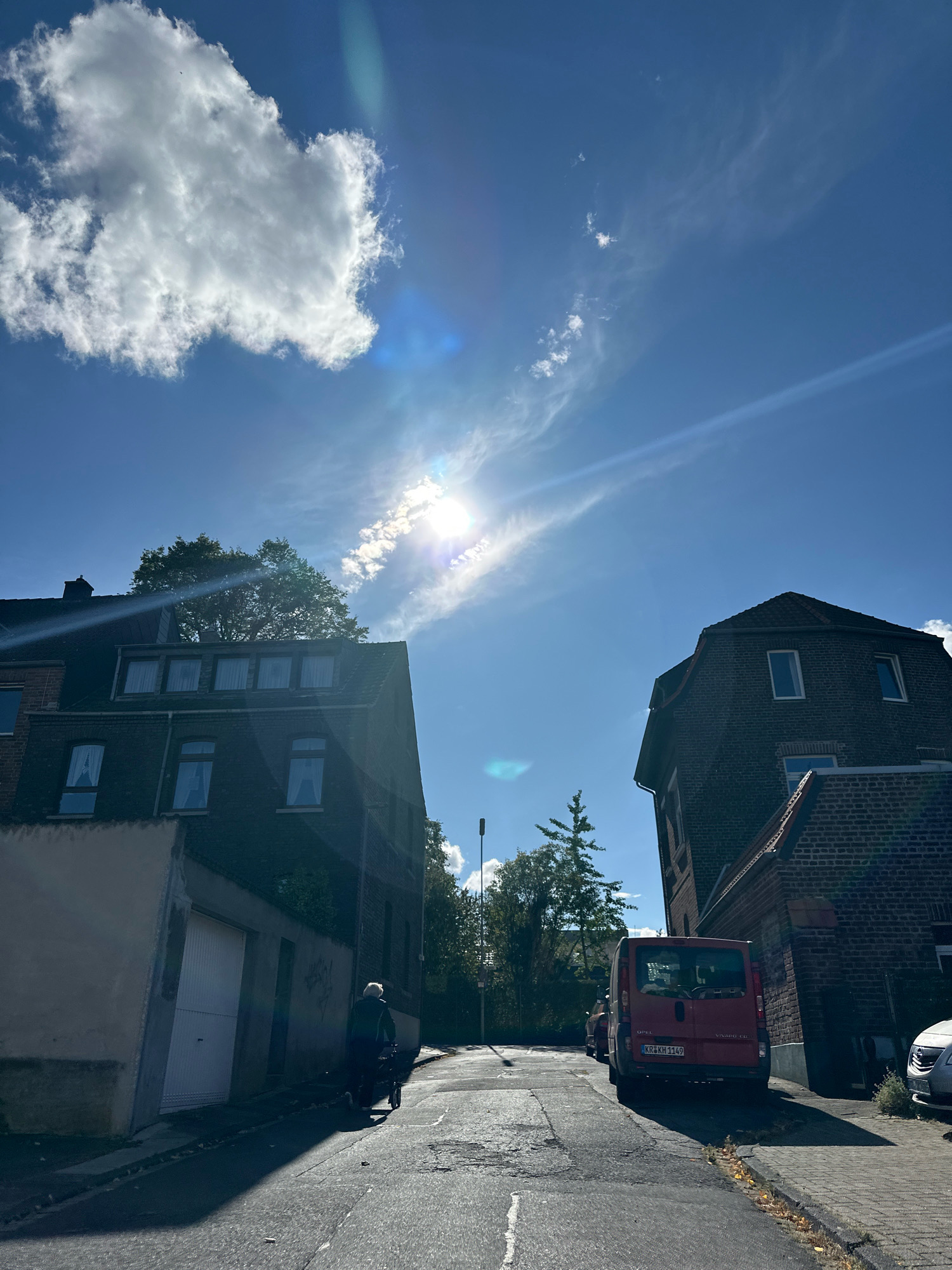 The image shows a street scene in Krefeld, Germany, on a sunny day with blue skies and scattered clouds. Two residential buildings, one on the left and one on the right, flank a narrow asphalt street. The building on the left is a dark brick structure with multiple windows and a sloped roof. The building on the right is also brick, with a steeply pitched roof and visible dormer windows. A red Opel Vivaro van is parked on the right side of the street, facing away from the camera. A person, seen from behind, is walking on the left side of the street, pushing a wheeled walker. The street is lined with sidewalks and patches of grass. The sun is high in the sky, casting long shadows.