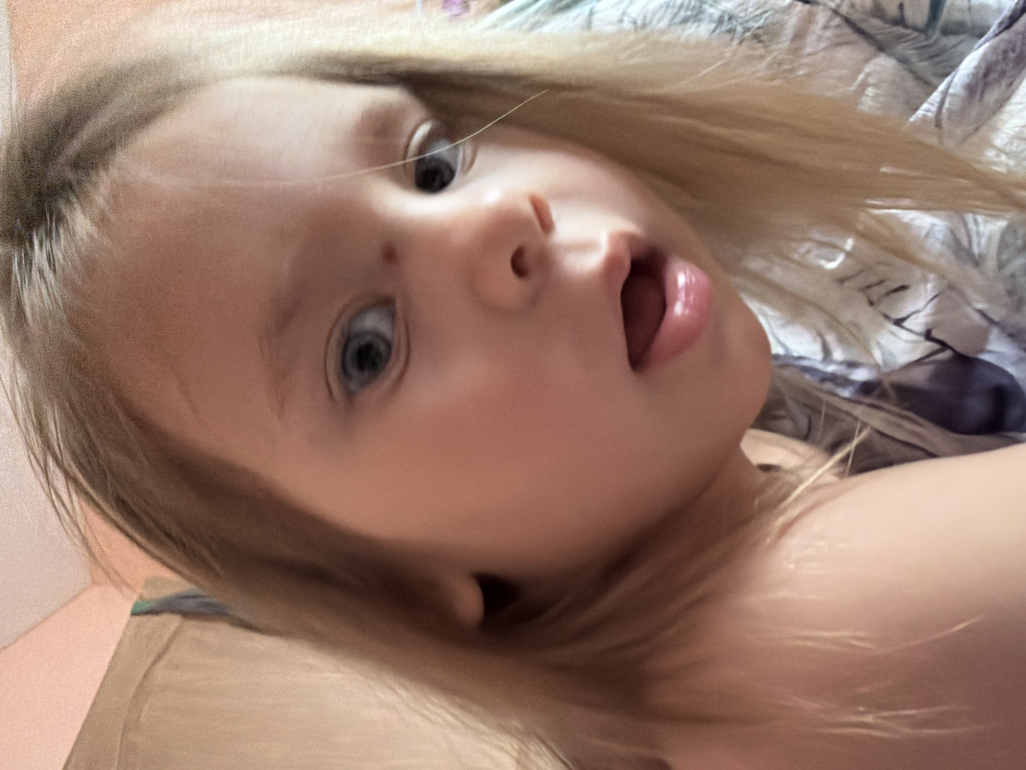 A close-up, diagonally oriented portrait features a young child with fair skin, light blonde hair, and wide blue eyes. The child's face is central to the frame, with an expression of surprise or curiosity indicated by their slightly open mouth, revealing their tongue, and wide eyes looking directly at the camera. Their blonde hair flows around their head and shoulders. The child appears to be lying down, possibly on a bed or sofa, with soft, light-colored fabrics visible beneath them and a patterned blue, gray, and white fabric in the upper right background. A portion of the child's bare shoulder is visible in the lower right. The lighting suggests an indoor setting during the daytime. The image captures a candid moment of the child in Chaikovsky, Russia. No text is visible.
