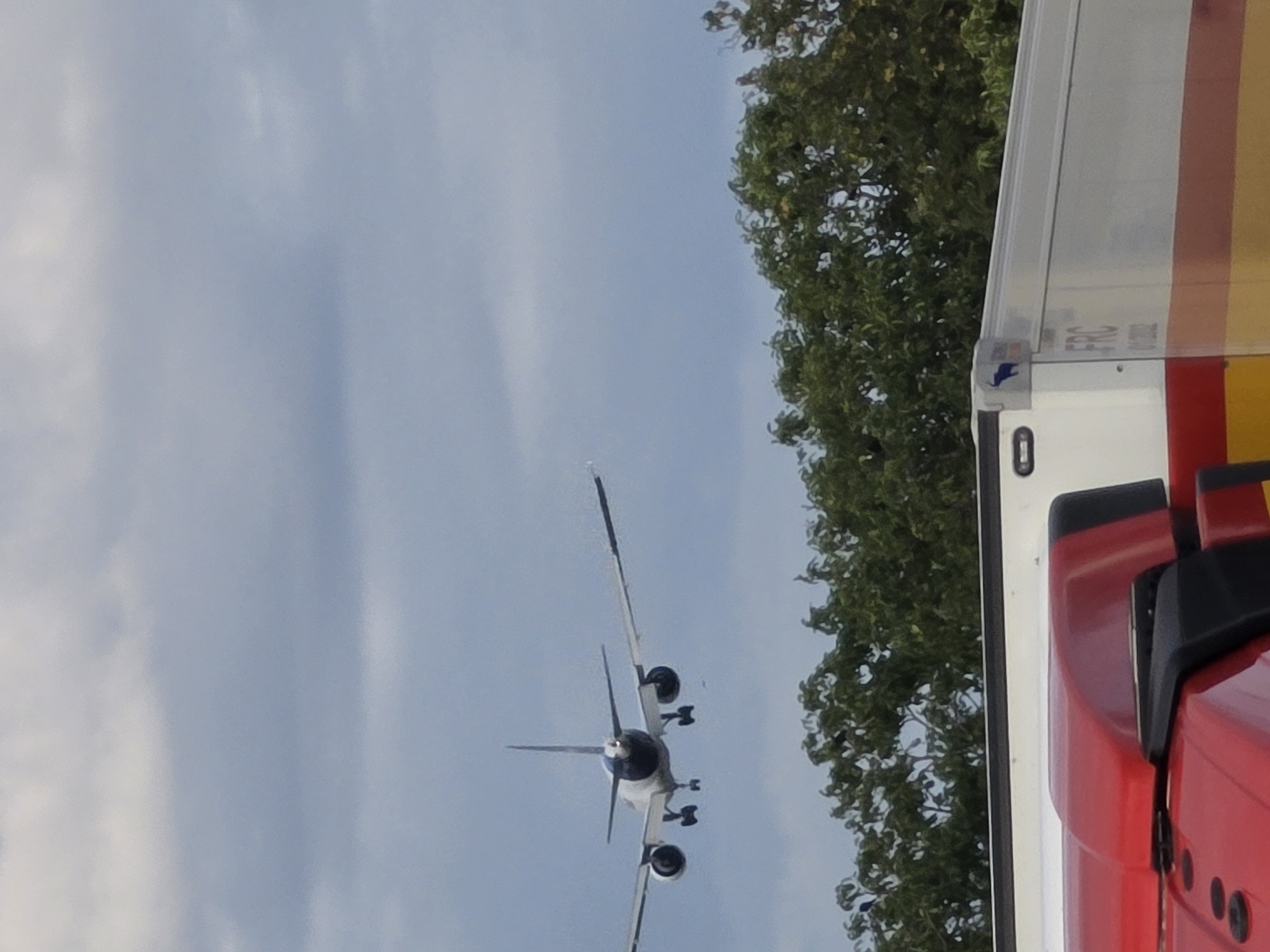 An airplane is in flight above a tree line, with its landing gear extended, suggesting it is preparing to land. Below the airplane, and partially obscuring the tree line, are the roofs of two vehicles, a red truck and a white trailer. The truck is in the foreground and appears to be a transport vehicle. The trailer is behind the truck, and has "FRC" printed on its side. The sky is overcast with light blue and white clouds. The image is taken from a low angle, looking upwards.