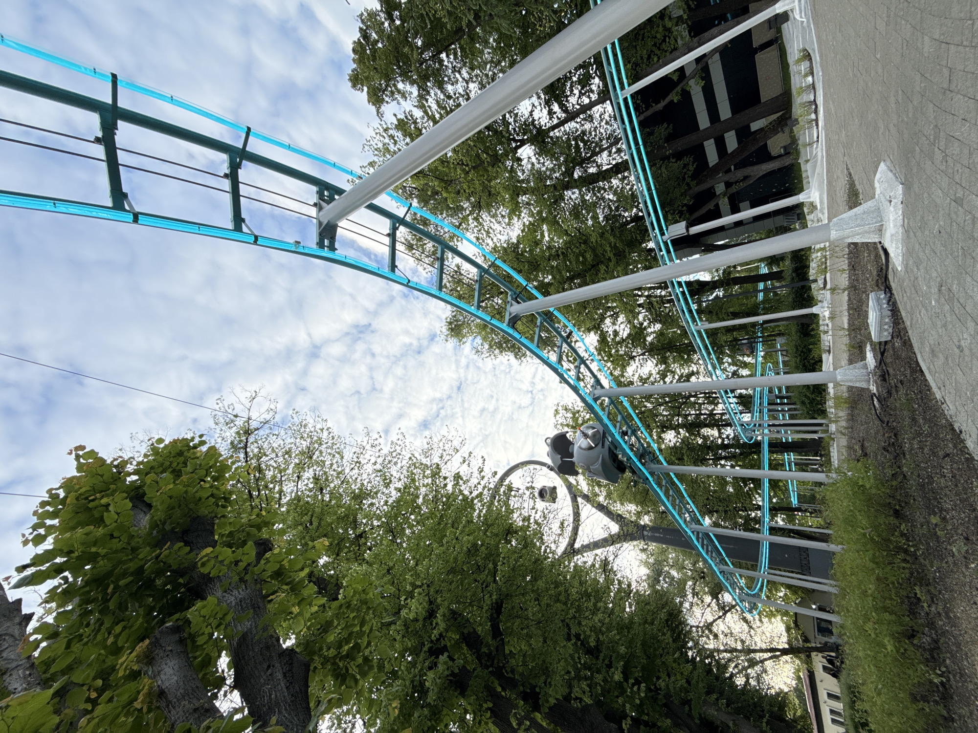 An outdoor, daytime shot captures a section of a light blue, illuminated amusement park ride track, likely a roller coaster or monorail, curving upwards through a verdant urban environment. The track, constructed from dark grey metal, features bright, translucent blue rails that appear lit, standing out against the cloudy sky. A single, empty grey gondola or seat is visible on the track, positioned mid-frame and moving upwards.
Lush green trees with dense foliage dominate the surroundings, framing the track on both sides and forming a vibrant backdrop. The sky above is bright with scattered white clouds, indicating clear daytime conditions. To the right, a paved walkway or sidewalk can be seen, bordered by low green bushes. In the background, beyond the trees, glimpses of dark-colored buildings with prominent windows are visible, suggesting an urban area. The illuminated track adds a striking, modern element to the natural setting. No people or discernible text are present. The scene depicts a recreational area, such as a park or amusement park, in Moscow, Russia.