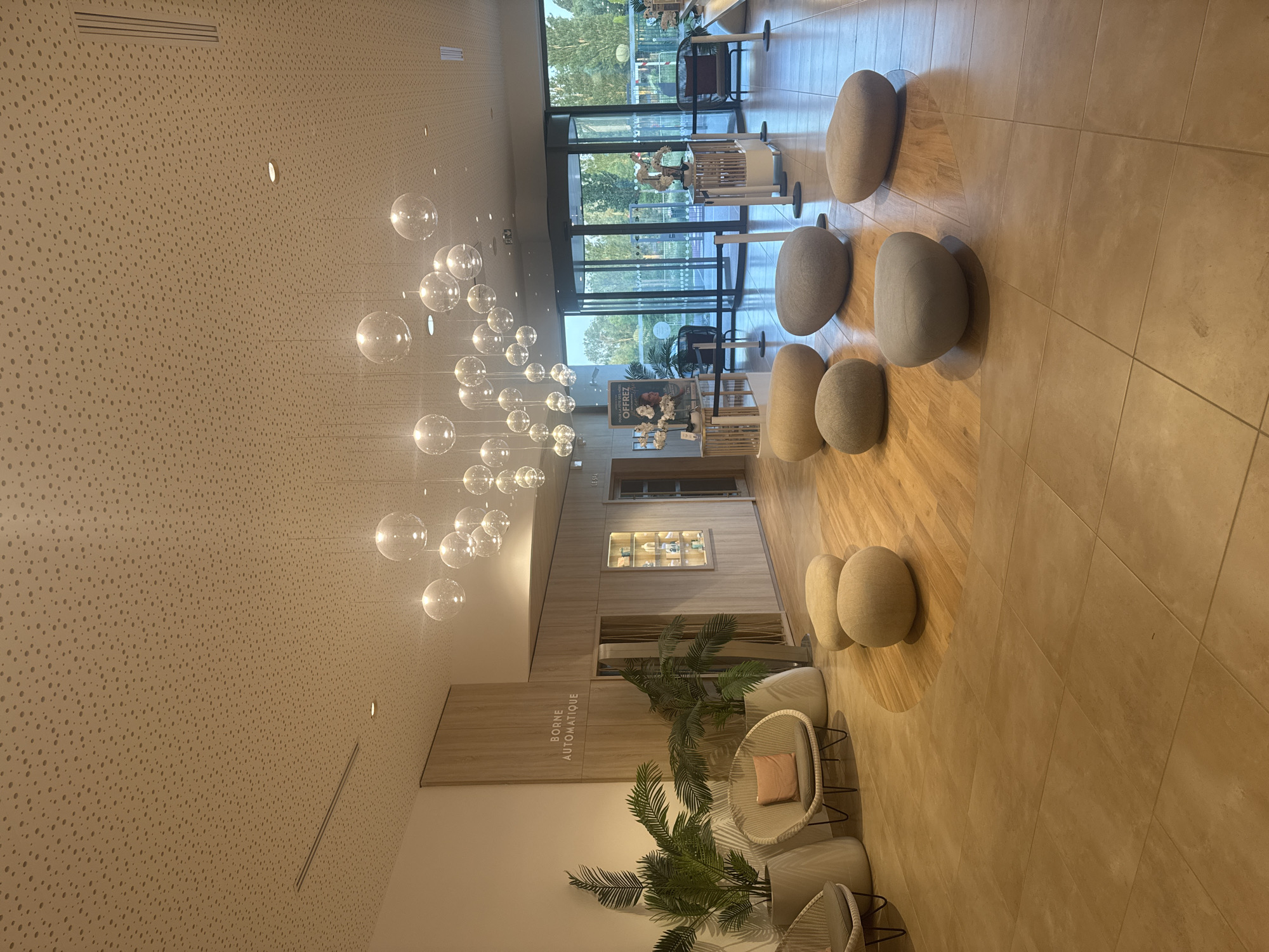 The image captures the interior of a modern building, located in Marcq-en-Barœul, France. The space features a polished tiled floor, leading towards large glass doors that offer an exterior view of greenery and a building across the street.
Overhead, a cluster of clear, spherical pendant lights hangs from the ceiling, creating a contemporary and airy atmosphere. The seating area is arranged with a series of pebble-like poufs in various shades of beige and gray. Wicker chairs with light-colored cushions and potted plants are also part of the seating arrangement.
To the side, a wooden structure with the words "BORNE AUTOMATIQUE" is visible, indicating the presence of an automated kiosk or service point. Further into the building, a display case filled with decorative objects can be seen.
The overall environment suggests a public space, such as a hotel lobby or a waiting area, designed with modern aesthetics and a focus on natural light and comfortable seating. There are no people visible in the image, and the setting appears calm and well-maintained.