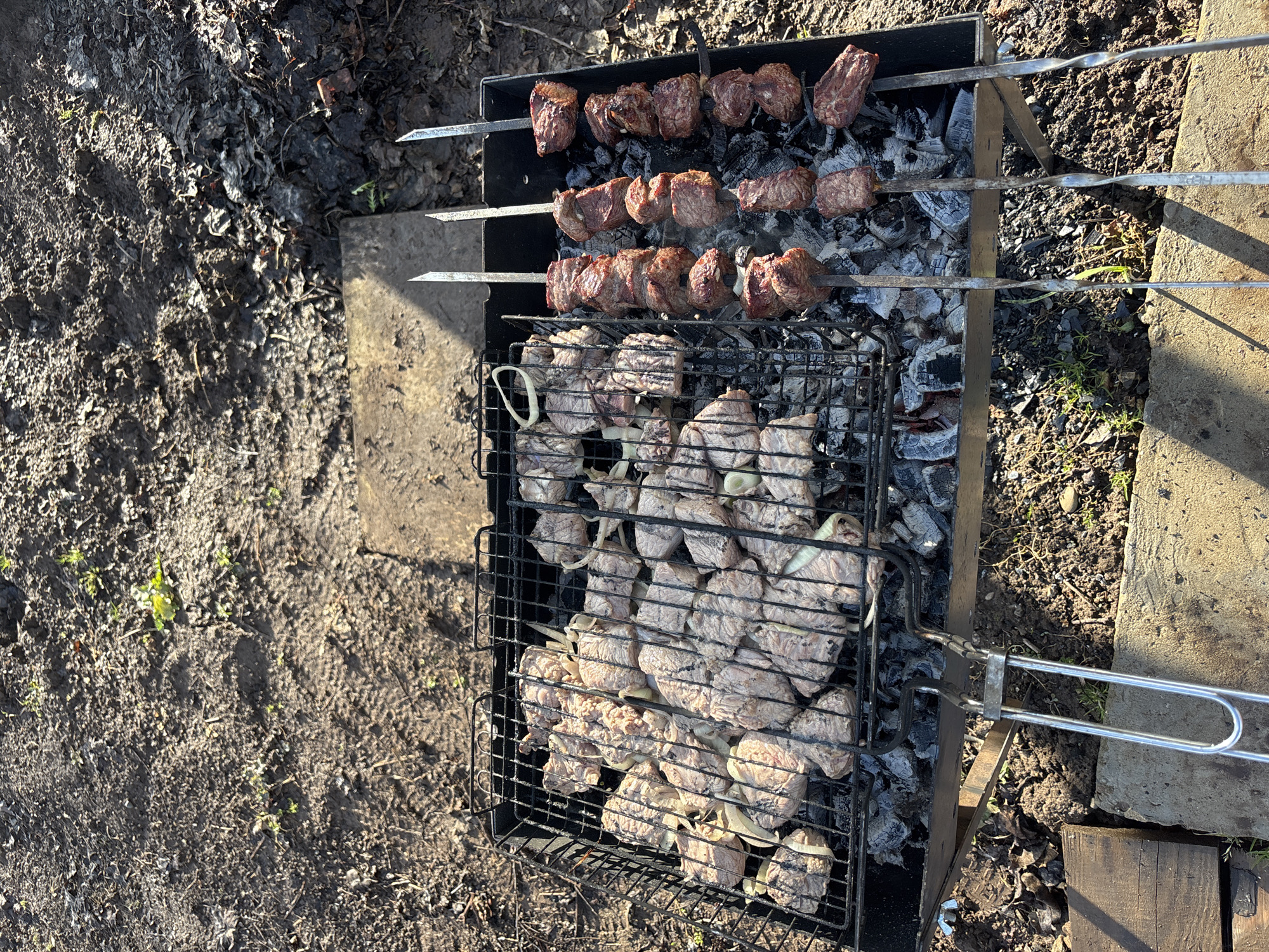A barbecue grill, located outdoors, contains partially cooked meat. Skewers with grilled meat are placed above a grill grate holding raw meat interspersed with onion slices. The grill stands on concrete slabs and wooden blocks, surrounded by dirt and leaf litter. The location is likely Chaikovsky, Russia.