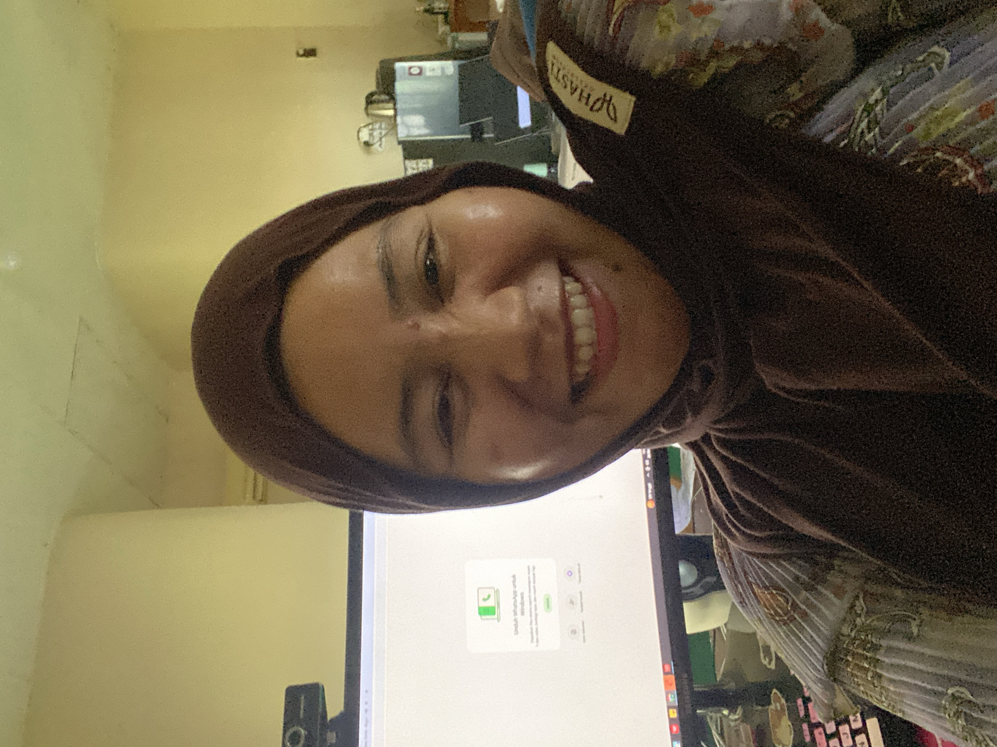 A medium shot captures a woman wearing a brown hijab and a patterned top, smiling at the camera. She is positioned in front of a computer monitor displaying a WhatsApp download page for Windows. A webcam is visible on top of the monitor. In the background, a refrigerator and other household items can be seen. The overall setting appears to be an indoor space, likely a home office, in Banjarbaru, Indonesia.