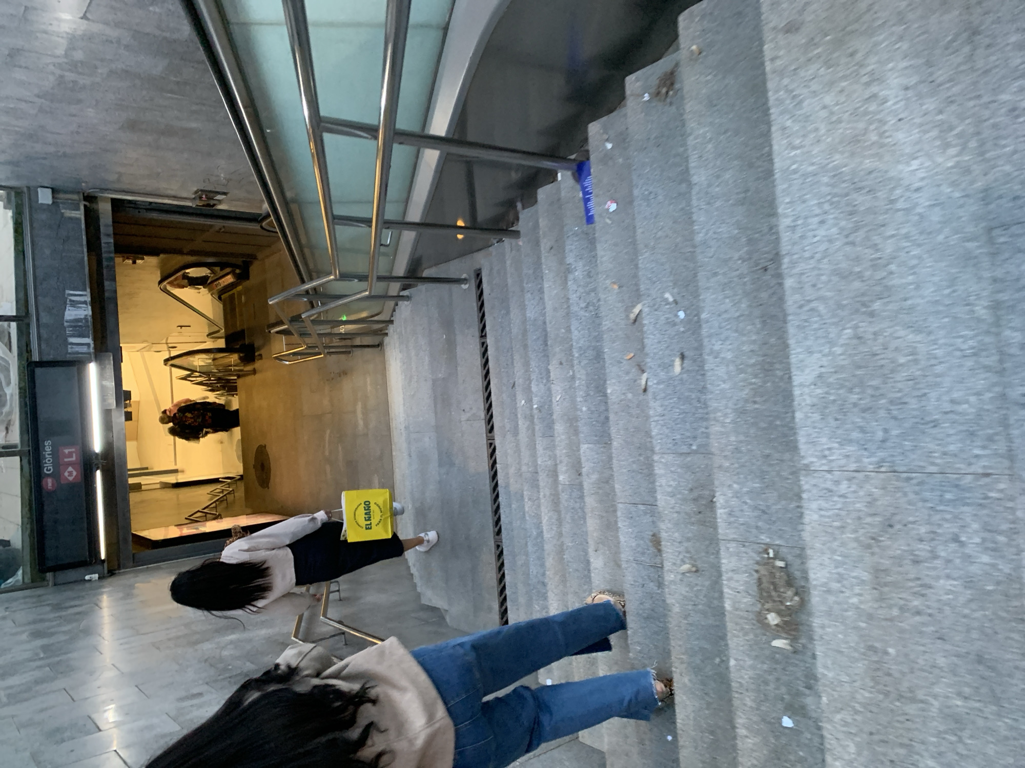 The image shows the Glories Metro station in Barcelona, Spain. The photo is taken from an elevated point looking down on a staircase leading to the station entrance. Two individuals are visible: One person is descending the stairs, wearing jeans and a light-colored jacket. The second person is standing near the station entrance, wearing dark pants and a light-colored top and carrying a yellow bag with "El Najro" printed on it. The Metro entrance is visible with signs "Glories L1". The staircase has metal handrails and a central drainage channel. The steps appear to be made of concrete.