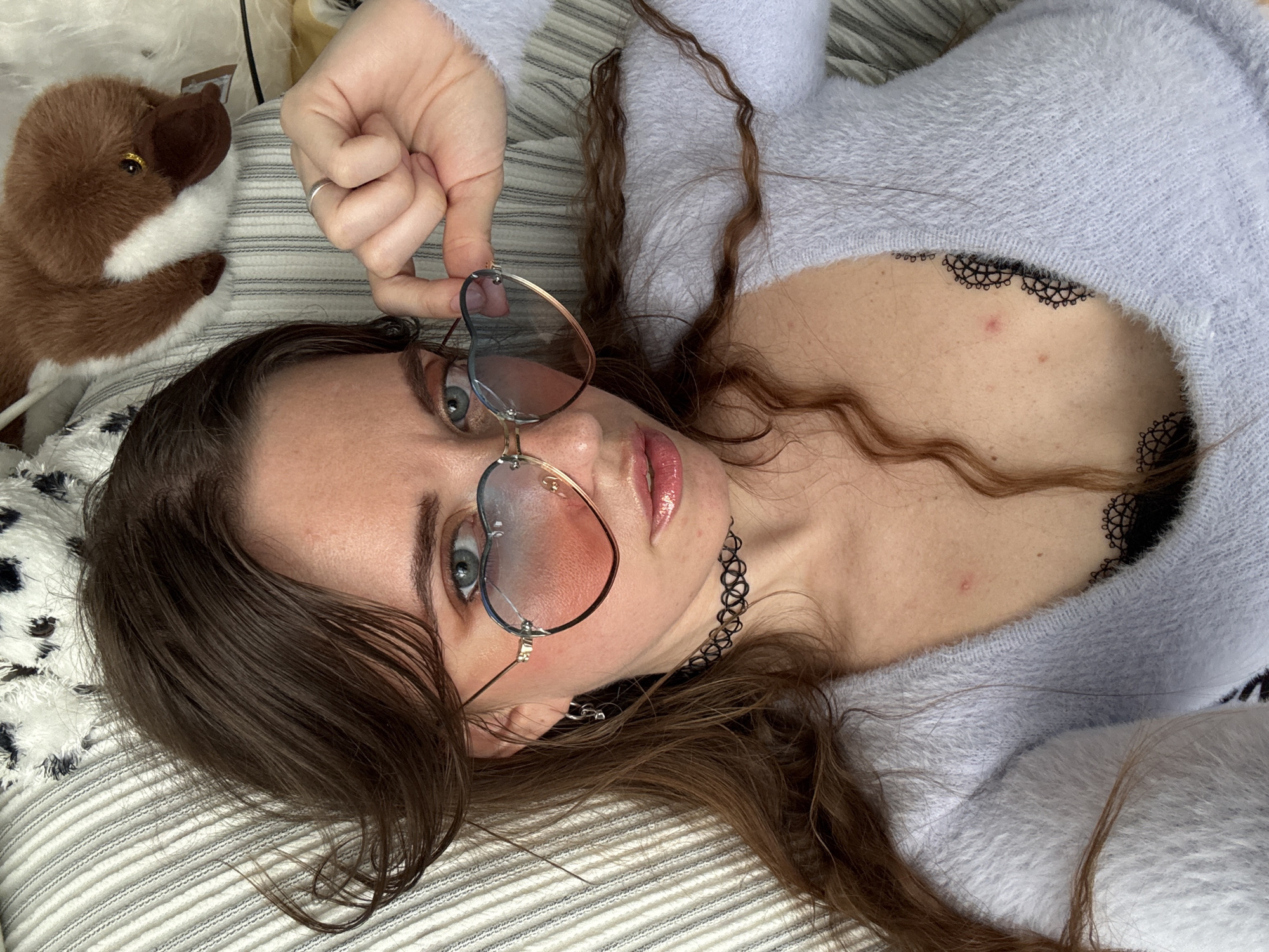 A young woman with long, dark brown hair is lying on a striped surface, possibly a bed or couch. She is wearing a light blue, fuzzy sweater and a black choker necklace. Her eyes are open and looking upwards. She is holding a pair of heart-shaped glasses with gold rims up to her face, obscuring part of her eyes. Her lips are painted with a glossy pink lipstick.
To the left of the woman, a small, brown stuffed animal with white accents and gold eyes is visible. The striped surface has a pattern of thin gray and white lines. The lighting suggests it is daytime, with no specific weather cues. The setting appears to be indoors, likely a bedroom or living space. There is no visible text in the image.