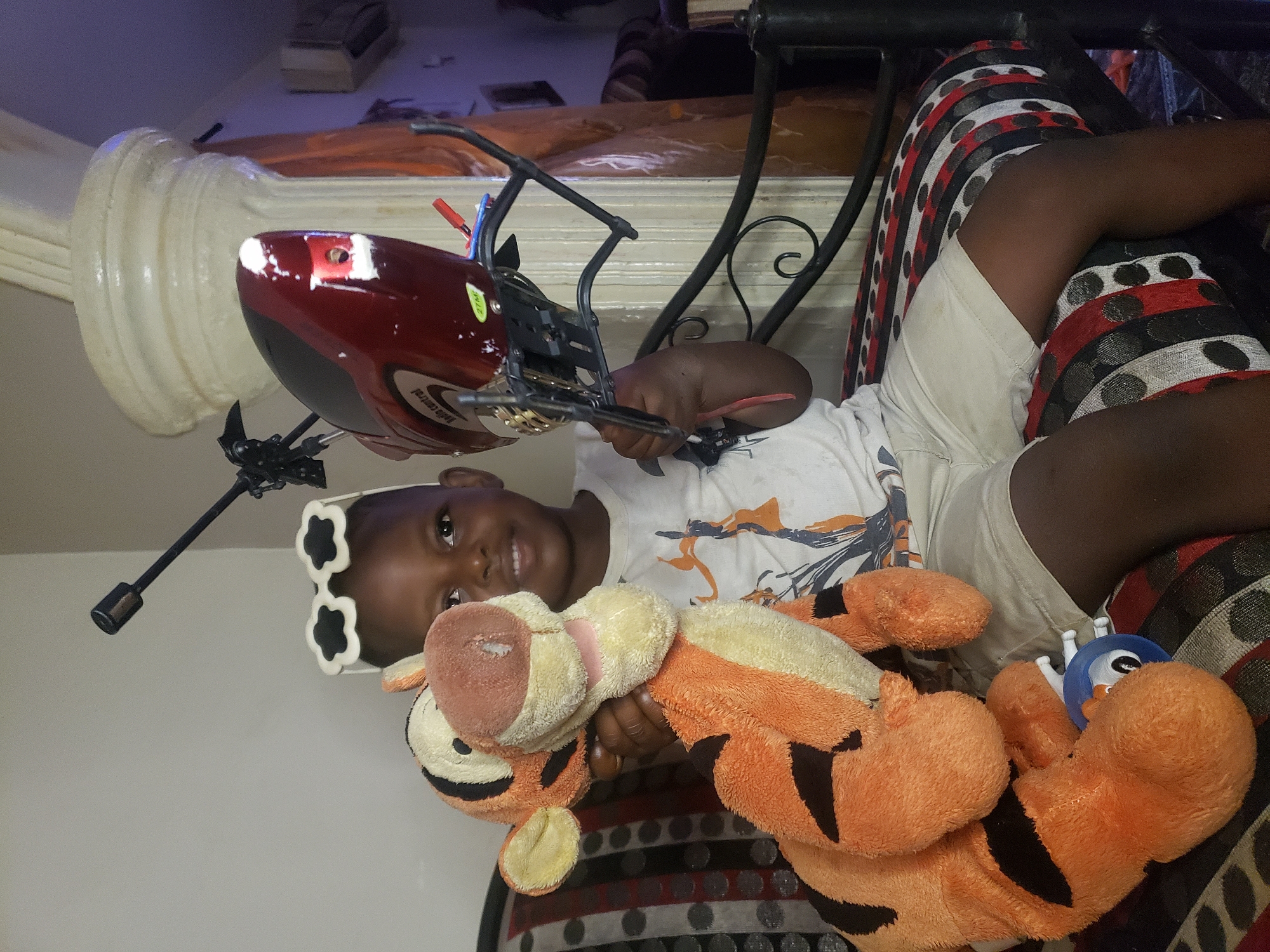 A young boy, appearing to be between 3 and 5 years old, sits on a patterned sofa in what is identified as Sabon Tasha, Nigeria. He is wearing a white t-shirt with a graphic print and light-colored shorts. On his head, he has novelty sunglasses with a cloud-shaped frame. In his right arm, he holds a large, stuffed toy tiger, and in his left hand, he clutches a red and black remote-controlled helicopter. The boy is smiling at the camera. Behind him, a large, fluted white column is visible, and part of a window with vertical blinds is also in the background. The lighting suggests an indoor setting, possibly a home environment, with artificial light sources contributing to the overall illumination. The sofa is upholstered with a fabric featuring a repeating pattern of black, red, and white circles and stripes.