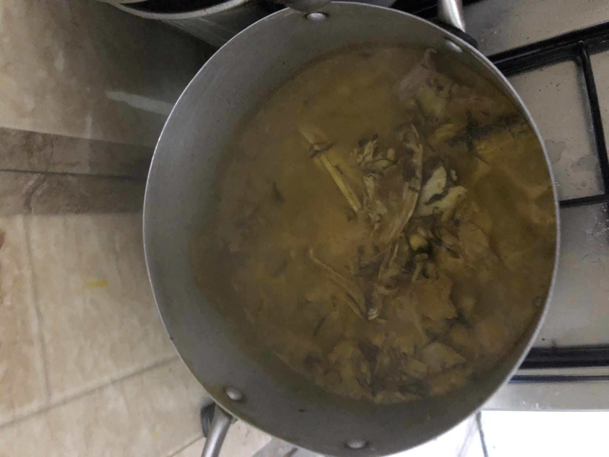 A pot of stew is cooking on a stovetop in a home kitchen in Kabong, Nigeria. The stew is a yellowish-brown color and appears to contain chunks of meat and bone. The pot is made of aluminum and has two handles. The stovetop is black and appears to be a gas stove. The background is tiled with light-colored tiles. There are no people visible in the image.