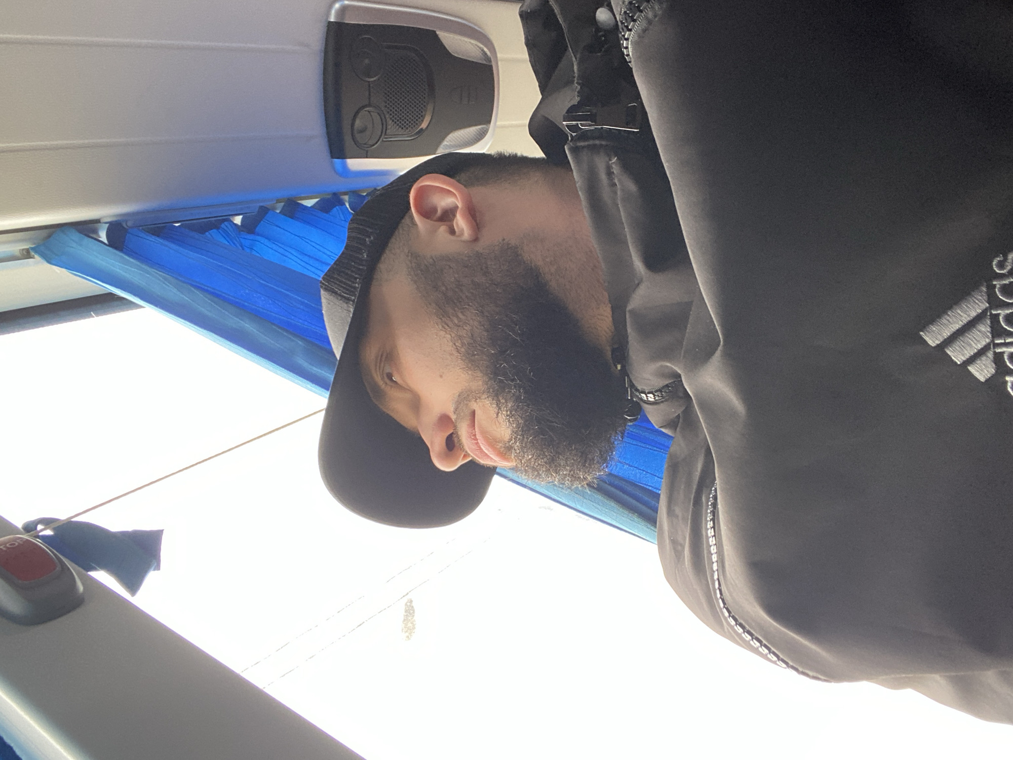 A medium shot depicts a man inside what appears to be a public transport vehicle, likely a bus or train, bathed in bright daylight. The man, seen from the mid-torso up, has a dark beard and mustache. He wears a black cap and a dark jacket featuring a white "Adidas" logo on the right side. His right ear is visible, and his gaze is directed forward or slightly downwards with a neutral expression.
The interior of the vehicle shows functional details: above the man's head, a ceiling-mounted panel contains a speaker grille and control knobs, characteristic of passenger seating. To his left, a blue pleated curtain or blind is partially drawn over a window, blocking some of the intense light. Further left and below, a white/grey panel with a prominent red "STOP" button and an attached cord confirms the setting as a public conveyance.
The exterior view through the window is overexposed, appearing as a bright white expanse, strongly suggesting direct sunlight and bright conditions, likely midday. The image's context is Kommunar, Russia.