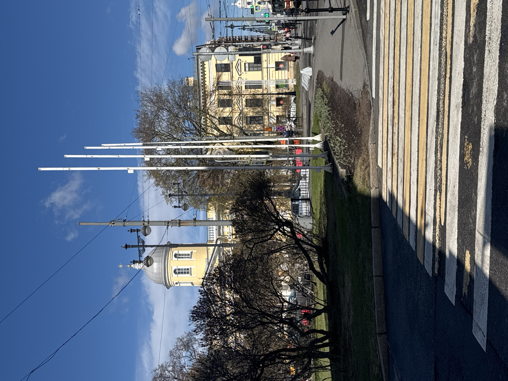The image displays an urban street scene in Saint Petersburg, Russia, under a bright blue sky with a few wispy clouds. The photograph appears to have been taken during daylight, likely mid-morning or afternoon, as evidenced by the distinct shadows cast across the pedestrian crossing.
A prominent, multi-story, light yellow building with classical architectural elements dominates the midground. Its facade features numerous rectangular windows, and a distinctive dome-like structure, topped with a cross or finial, is visible on its left side. Several red, square-shaped logos, too indistinct to read, are affixed to the building's exterior, potentially indicating businesses.
In the foreground and midground on the right, a wide pedestrian crossing with alternating white and yellow stripes spans the asphalt road. Adjacent to the road, a narrow strip of green grass with some sparse, low-lying vegetation runs alongside the curb.
Several trees with mostly bare branches, suggesting late winter or early spring, are scattered throughout the scene. One large, dark tree stands in the left foreground, partially obscuring the view of the yellow building. Numerous utility poles, streetlights, and a dense network of overhead wires crisscross the sky, characteristic of a city street. In the distance, several blurry figures of people can be seen walking on the sidewalk near the yellow building, and parts of parked vehicles are visible further down the street. No discernible text is clearly visible on any signs or objects.