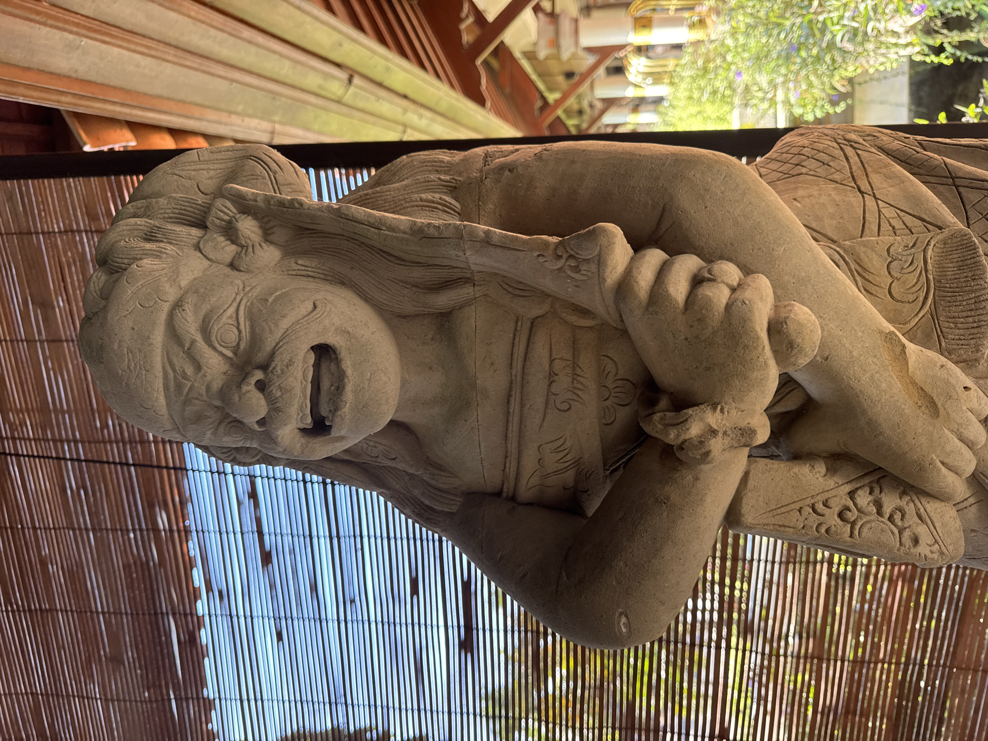 A light-colored stone statue of a mythological guardian figure stands prominently in the foreground. The figure features a fierce expression with large eyes, a prominent mustache, and an open mouth revealing teeth. It wears a carved headdress or elaborate hair and an ornate garment with a chest band and patterned lower covering. The figure holds a short, curved sword or dagger in its hands, positioned across its chest. To the left, a bamboo slat blind covers an opening, through which a bright blue sky and distant green foliage are visible. To the right, architectural elements of a building, including wooden ceiling beams and indistinct white columns with surrounding green plants, are present. The scene is brightly lit by natural daylight in Kuta Selatan, Indonesia.