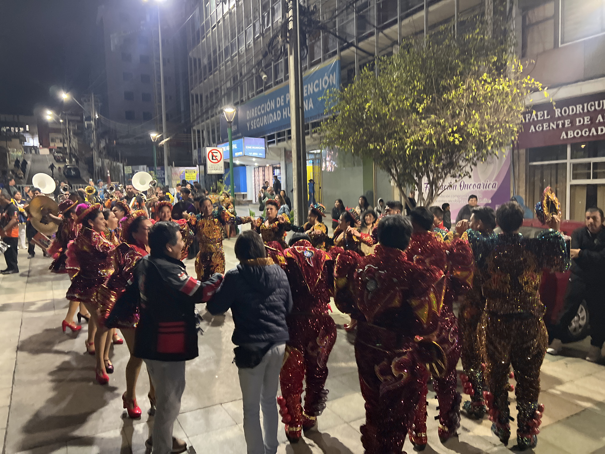 An evening street parade is depicted in Arica, Chile, featuring a group of performers in ornate, sequined costumes. The individuals are adorned in vibrant reds, golds, and blues, with elaborate headdresses and masks. Some performers are playing musical instruments, including large brass horns, suggesting a musical procession. The scene takes place on a paved street lined with buildings. A banner is visible on one building, with text in Spanish that appears to be part of a government or institutional announcement. A red circular sign with a white "E" is also present, indicating parking regulations. The overall atmosphere suggests a cultural event or celebration.
