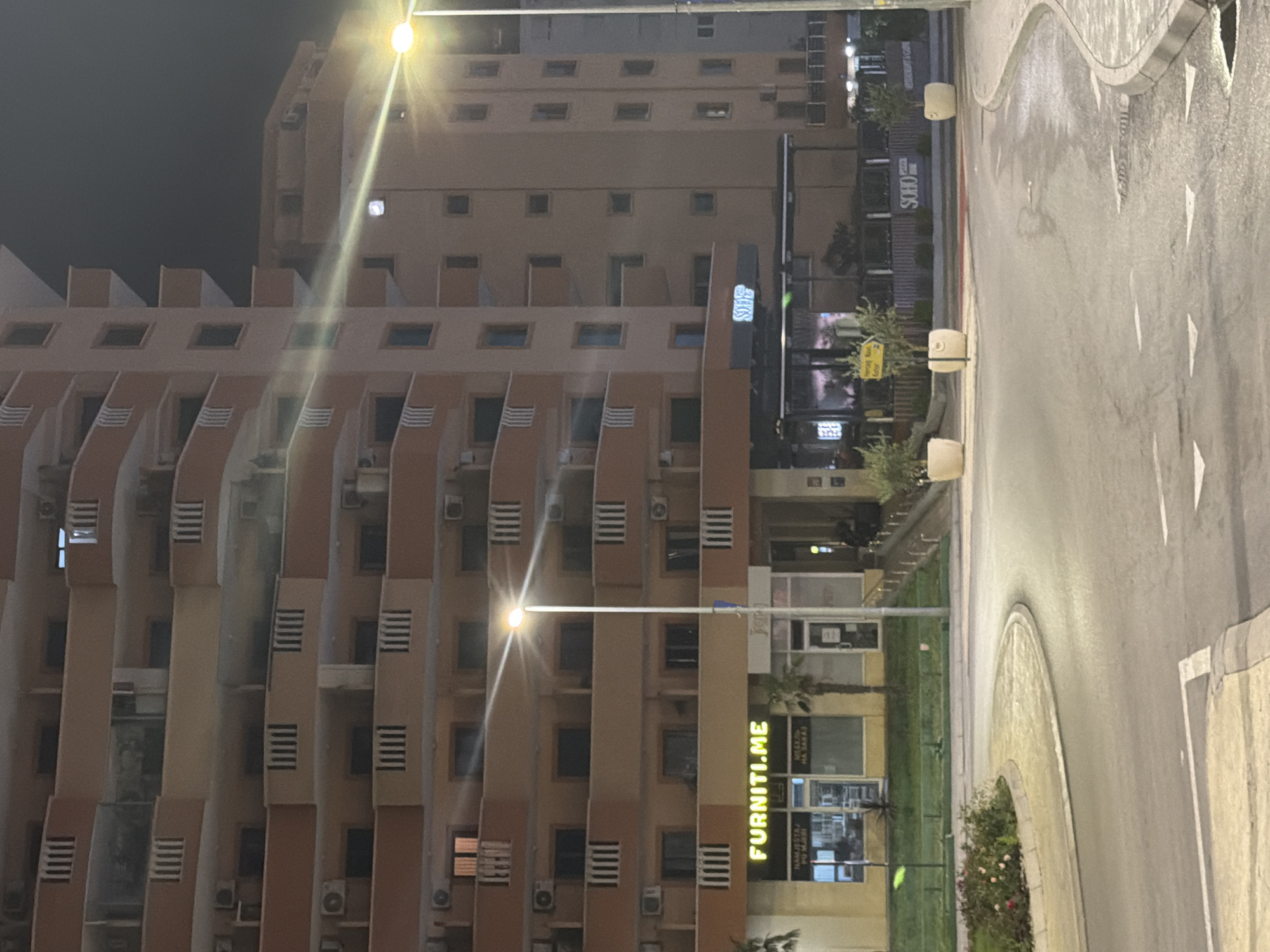 The image depicts a nighttime scene on a street in Budva, Montenegro. In the foreground, a wet asphalt road curves to the right, with white directional arrows painted on its surface. A roundabout with a circular flower bed containing red and pink roses is visible on the left. In the midground, a modern multi-story building dominates the scene, its facade a light orange-brown color with repeating balconies. Several shops are located at the ground level of the building. One shop displays a neon yellow sign that reads "FURNITI.ME." Another shop has a sign with "SOHO PIZZA." A street lamp with a bright light, creating a lens flare effect, stands prominently in the middle of the image. The background is dark, suggesting a clear night sky. The overall impression is of a commercial and residential area at night, with the street lights and building illumination providing the primary light sources.