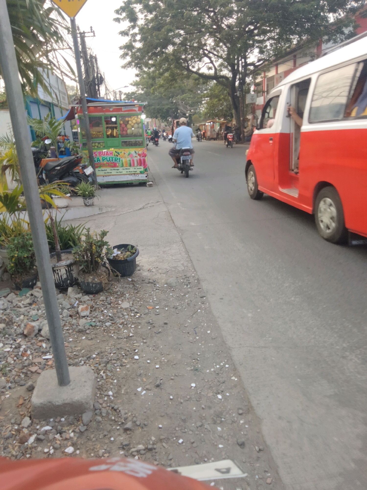 In Sepatan, Indonesia, a red and white van travels on a paved road alongside a fruit cart. A man on a motorcycle rides past the cart, which displays various fruits and beverages. The scene appears to be an outdoor market or roadside, with trees and buildings lining the street. The ground is covered with gravel and debris.