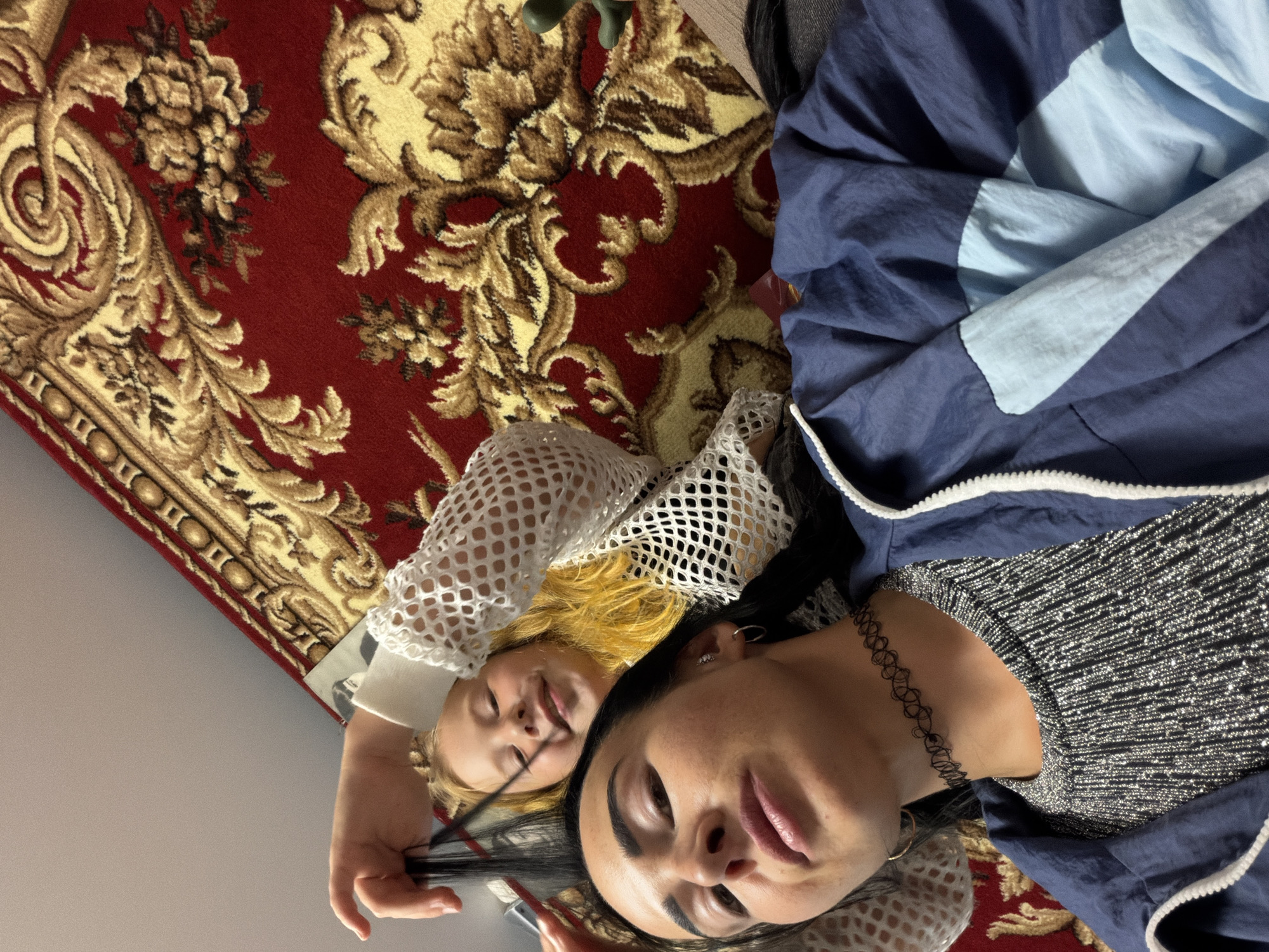 Two people, an adult and a child, are lying down on a red patterned rug. The adult has dark hair and is wearing a sparkly silver top and a blue jacket with light blue accents. They are looking at the camera. The child has blonde hair and is wearing a white mesh top. The child is smiling and looking towards the adult. The scene appears to be indoors, as suggested by the rug and clothing. The lighting is bright, suggesting it could be daytime. There is no visible text in the image. The overall mood is casual and intimate, possibly a candid moment between the two individuals. The backdrop of the patterned rug adds a touch of traditional decor to the scene.