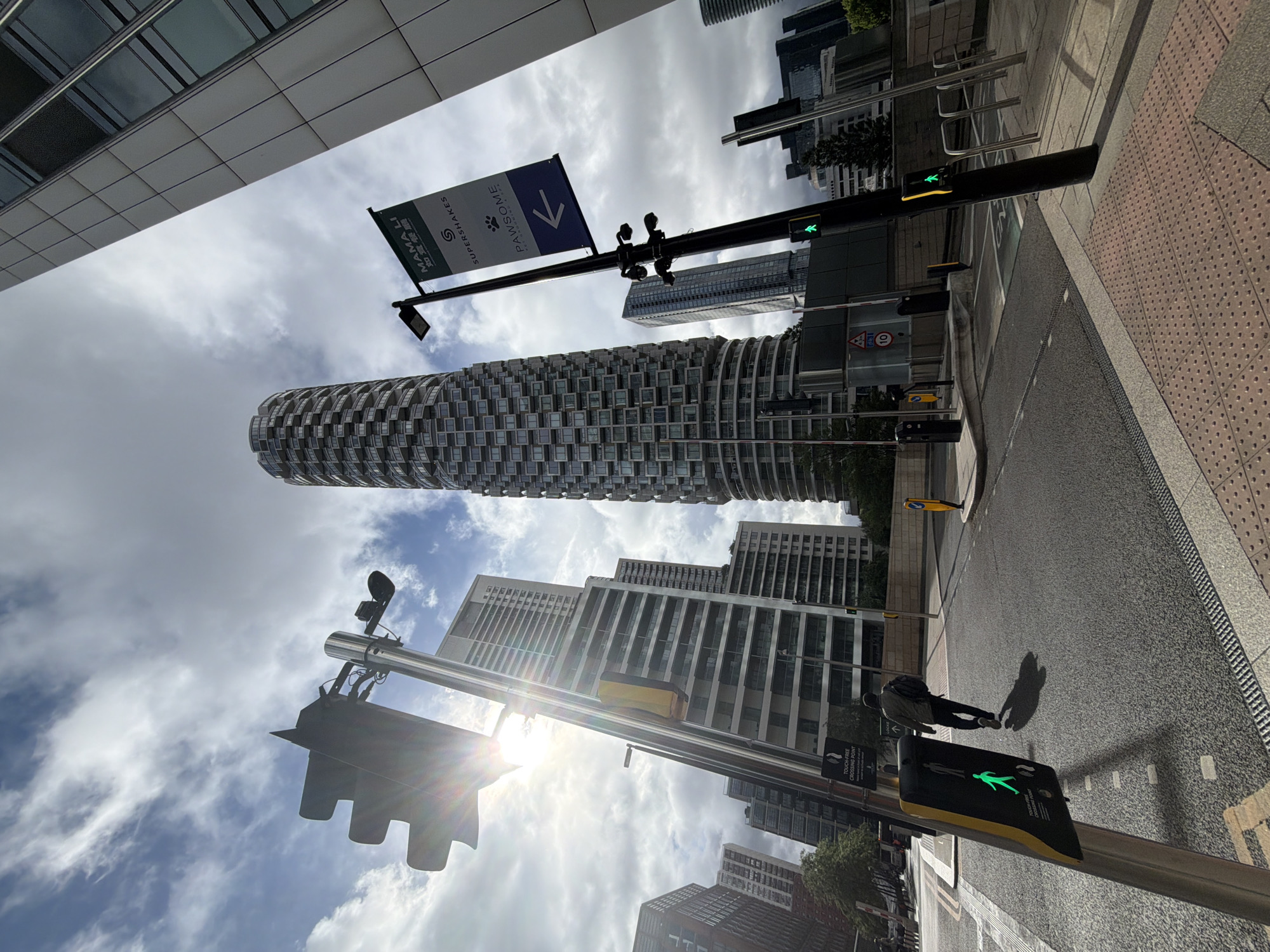 The image depicts a vibrant city street scene during daytime, under a partly cloudy sky with bright sunshine. The perspective is looking upwards from street level, highlighting towering modern architecture.
A prominent, cylindrical high-rise building with a distinctive patterned facade of windows and balconies stands centrally, its unique design catching the eye. Several other contemporary high-rise buildings with glass facades are visible in the background, framing the street.
In the foreground, a paved street with a textured tactile paving strip on the sidewalk is visible on the right. A black street light pole with a green pedestrian 'walk' signal is positioned on the right. Another traffic light further down the street also displays a green signal. The sun, located to the left side of the frame, casts a strong lens flare across the upper left portion of the image and illuminates the scene brightly, creating defined shadows.
A person, viewed from behind, is walking on the sidewalk, carrying a backpack and casting a long shadow ahead of them.
Visible text on a blue directional sign banner, attached to a street pole, displays white text reading "MAMALI", "Supershakes", and "PAWSOME" (accompanied by a paw print logo), along with a right-pointing arrow. Beneath "MAMALI", small, possibly Chinese characters are also present. On the pedestrian signal posts, the text "TOUCH-FREE CROSSING POINT" is clearly visible, and a glimpse of "PRESS BUTTON TO CROSS" can be seen on another sign. A small circular road sign with the number "10" is also present, likely indicating a speed limit.
The overall scene depicts a modern, bustling urban environment.