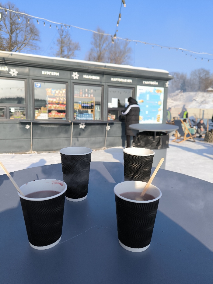 The image displays an outdoor winter scene in Prybuzke, Ukraine, under a clear blue sky with string lights overhead. In the foreground, a dark grey circular table holds four black, ripple-textured paper cups with white rims. Three cups contain a steaming, dark red liquid; one of these includes a light-colored wooden stirring stick. The fourth cup also emits steam but appears empty. In the midground, a dark-colored rectangular kiosk with multiple windows features Cyrillic signage above, identifying offerings such as 'TEA,' 'BURGERS,' 'DRINKS,' 'FRENCH FRIES,' and 'GLÜHWEIN.' A lone individual, dressed in a dark winter coat with white arm stripes, stands at a kiosk service window. Adjacent to the kiosk, a dark round outdoor table and a black cylindrical trash bin are present. The ground is uniformly covered in snow. In the background, deciduous trees are visible without foliage, alongside a snow-covered bank where several indistinct figures are observed, some seated in wooden leisure chairs.