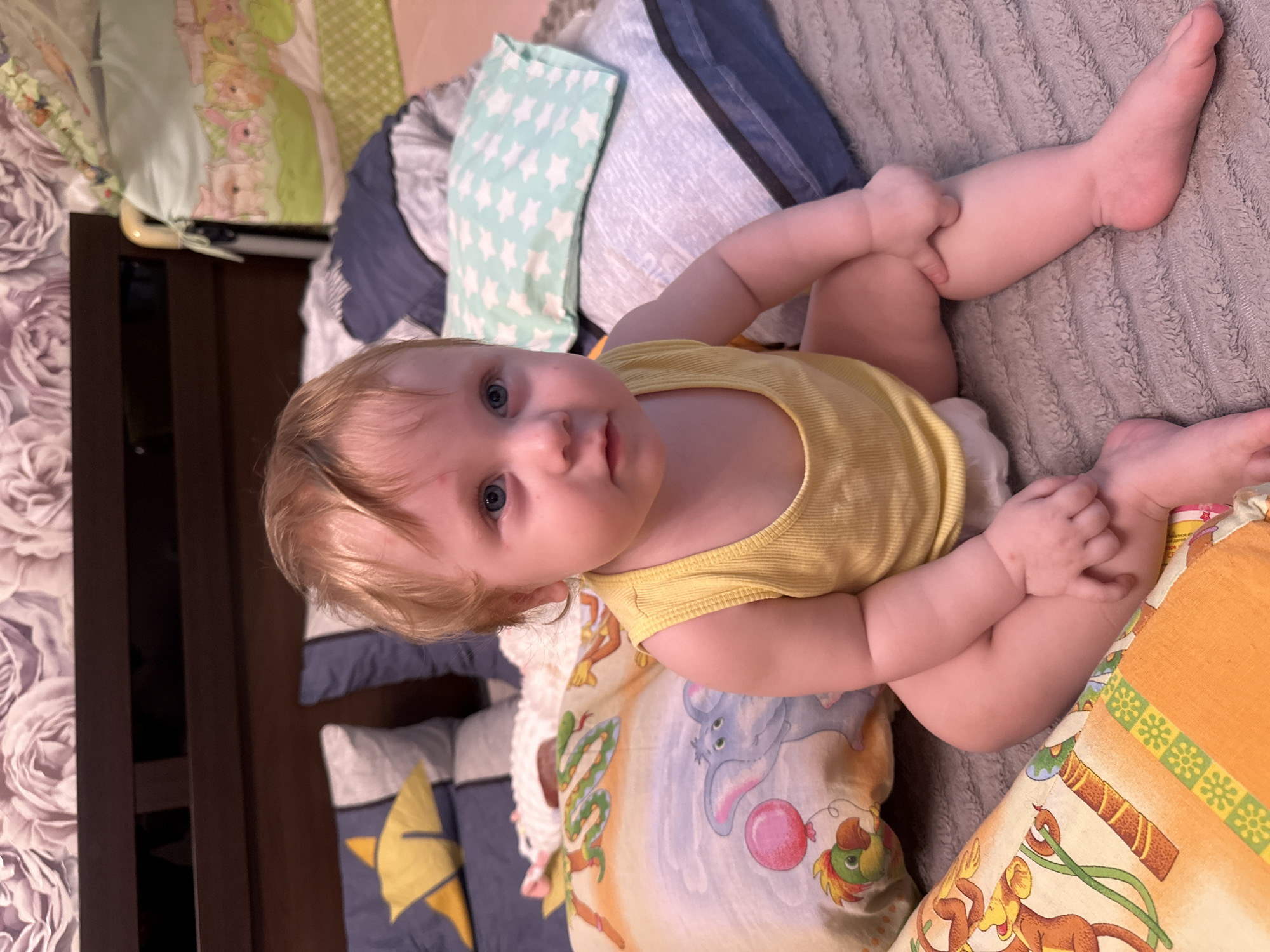An infant with blonde hair and blue eyes is sitting on a bed. The infant is wearing a yellow tank top and a diaper. The infant is surrounded by pillows with various patterns, including stars, elephants, and monkeys. The background shows a headboard and a crib. The image appears to be taken indoors, likely in a bedroom. The location in Sayanogorsk, Russia is unknown and cannot be determined from the image alone.