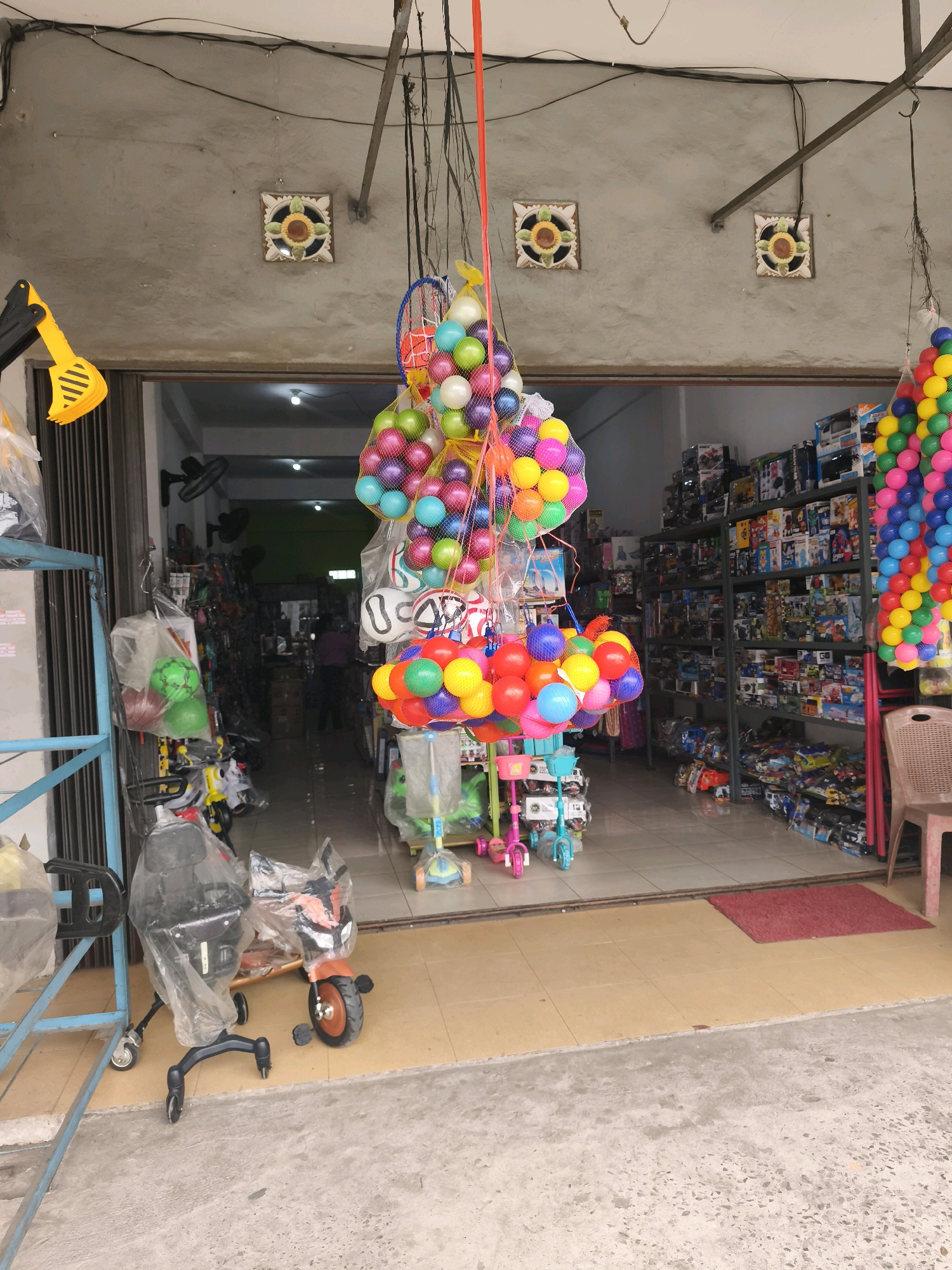 A roadside toy store in Palembang, Indonesia, displays a variety of children's toys and recreational items. The store appears to be situated in an open-air setting with a concrete facade and an open storefront. Inside, shelves are stocked with boxed toys, and various items are displayed outside, including inflatable balls in netting, a bicycle, and a miniature motorcycle covered in plastic wrap. The scene is brightly lit, suggesting it is daytime. The store is likely a small business catering to local residents and tourists.