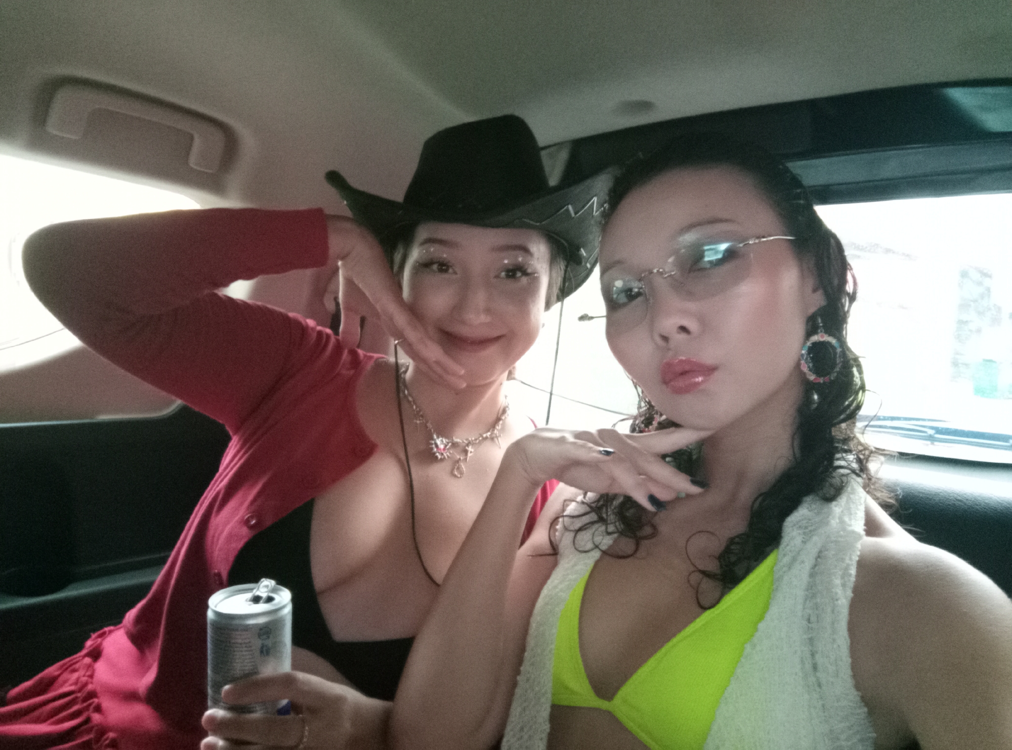 Two women are posing for a selfie in the back seat of a car. The woman on the left is wearing a black cowboy hat, a red cardigan over a black top, and a necklace. She is holding a silver can, possibly a beverage. The woman on the right is wearing glasses, a neon green bikini top, and has a white towel draped around her neck. Both women are touching their faces with their hands. The interior of the car is visible, with a window and a grab handle. The location is likely in North Kuta, Indonesia.