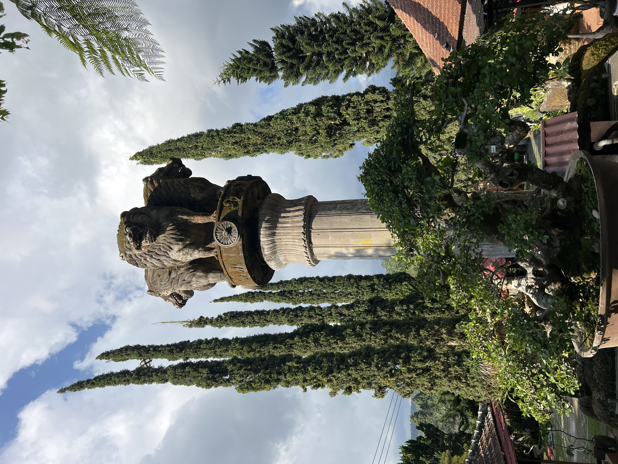 The image shows a stone monument topped with four lion heads, reminiscent of the Ashoka pillar, set against a backdrop of tall cypress trees and a cloudy sky. In the foreground, a large, meticulously pruned bonsai tree dominates the lower half of the frame. The scene appears to be in a garden or park-like setting, with tiled roofs of buildings visible in the middle ground and lush greenery in the distance. The lighting suggests it is daytime, with some areas illuminated by sunlight and others in shadow. The monument and bonsai tree suggest a cultural or aesthetic interest, possibly a tourist attraction or a private collection in Pupuan, Indonesia. There are no people visible in the image.