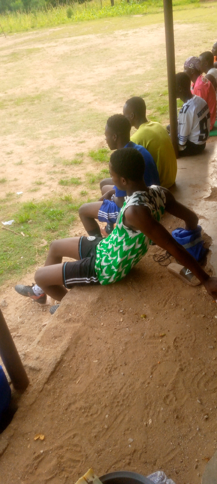 The image shows a group of young people sitting on a raised dirt platform next to a concrete structure, possibly a pavilion or bleachers, in Langtang, Nigeria. The foreground is dominated by the dirt platform and the people sitting on it. The person in the immediate foreground, facing away from the camera, is a young person wearing a green and white patterned sleeveless shirt and black shorts with white stripes. They are leaning back on their hands, with their legs extended. To their left, another young person is visible, wearing a blue shirt and black shorts with white stripes. Further back, other individuals are seated, some wearing t-shirts of various colors, including yellow and white with black numbering. The background consists of a grassy field with some vegetation and trees. The overall setting appears to be an outdoor recreational area, likely a sports field or training ground. The lighting suggests it is daytime.