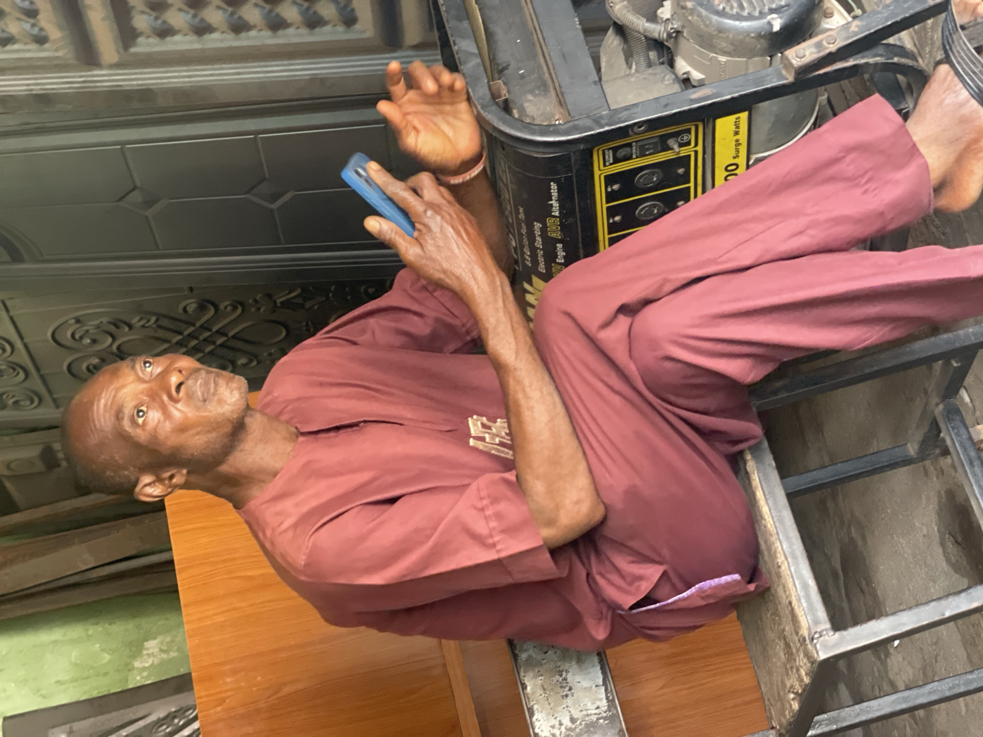A senior man is sitting sideways on a metal shelf, adjacent to a black generator. He holds a blue phone. He is wearing a brown shirt and trousers. The setting appears to be inside or adjacent to a building with a decorated black door. The image was taken in the city of Zaria, Nigeria.