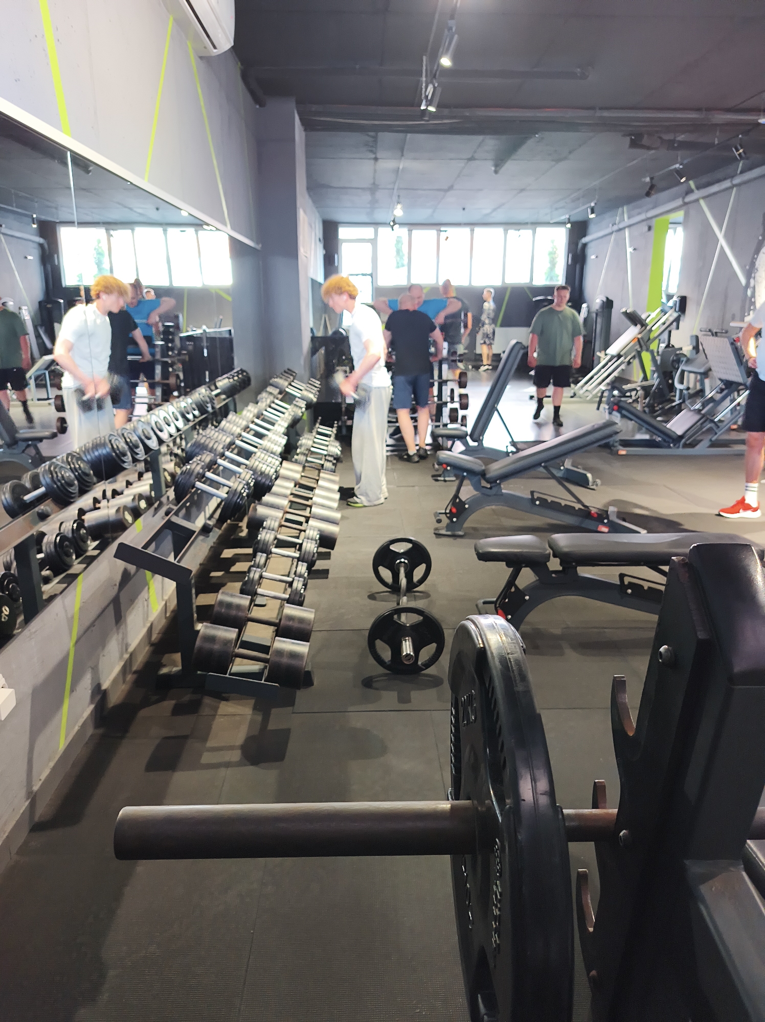 The image depicts the interior of a gym, likely in Ternopil, Ukraine, given the context. A mirrored wall runs along the left side, reflecting the gym's contents. Rows of dumbbells are arranged in a gray metal rack. Several people are present, engaged in various activities. One individual is seen holding a dumbbell, while others are standing near weight machines. Weight benches are scattered throughout the space. Additional weights, including plates, are visible on the floor. The flooring is black rubber. The gym has a high ceiling, with visible lighting fixtures.